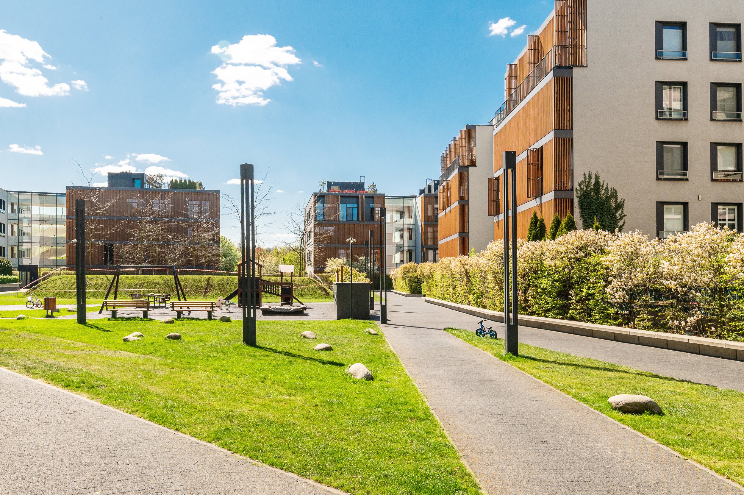 Residential apartment complex with a landscaped courtyard, playground, walking paths, benches, and bicycles on a sunny day.