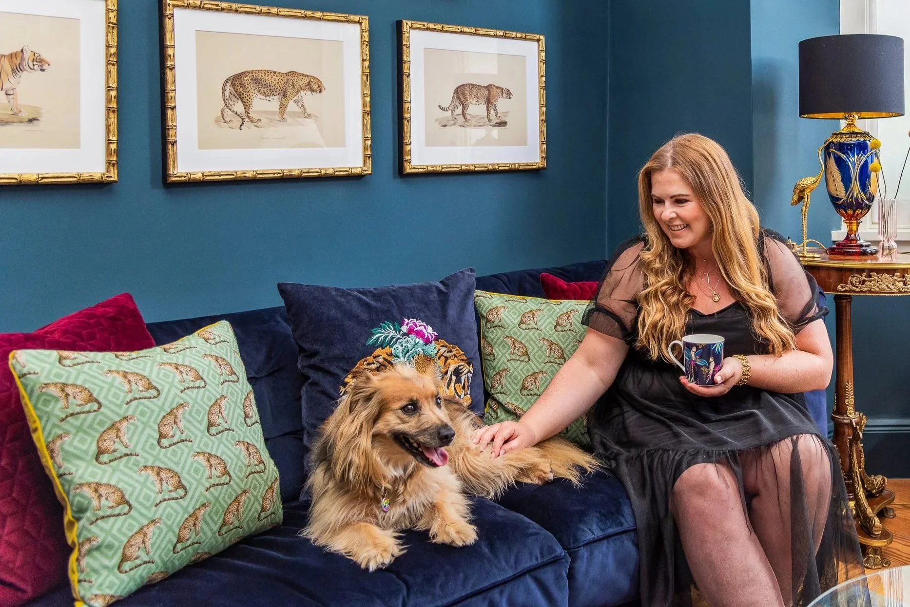 A woman sitting on a blue couch with a golden retriever puppy, surrounded by pillows with jungle animal patterns, in a room with framed pictures of big cats on the wall and exotic decor.