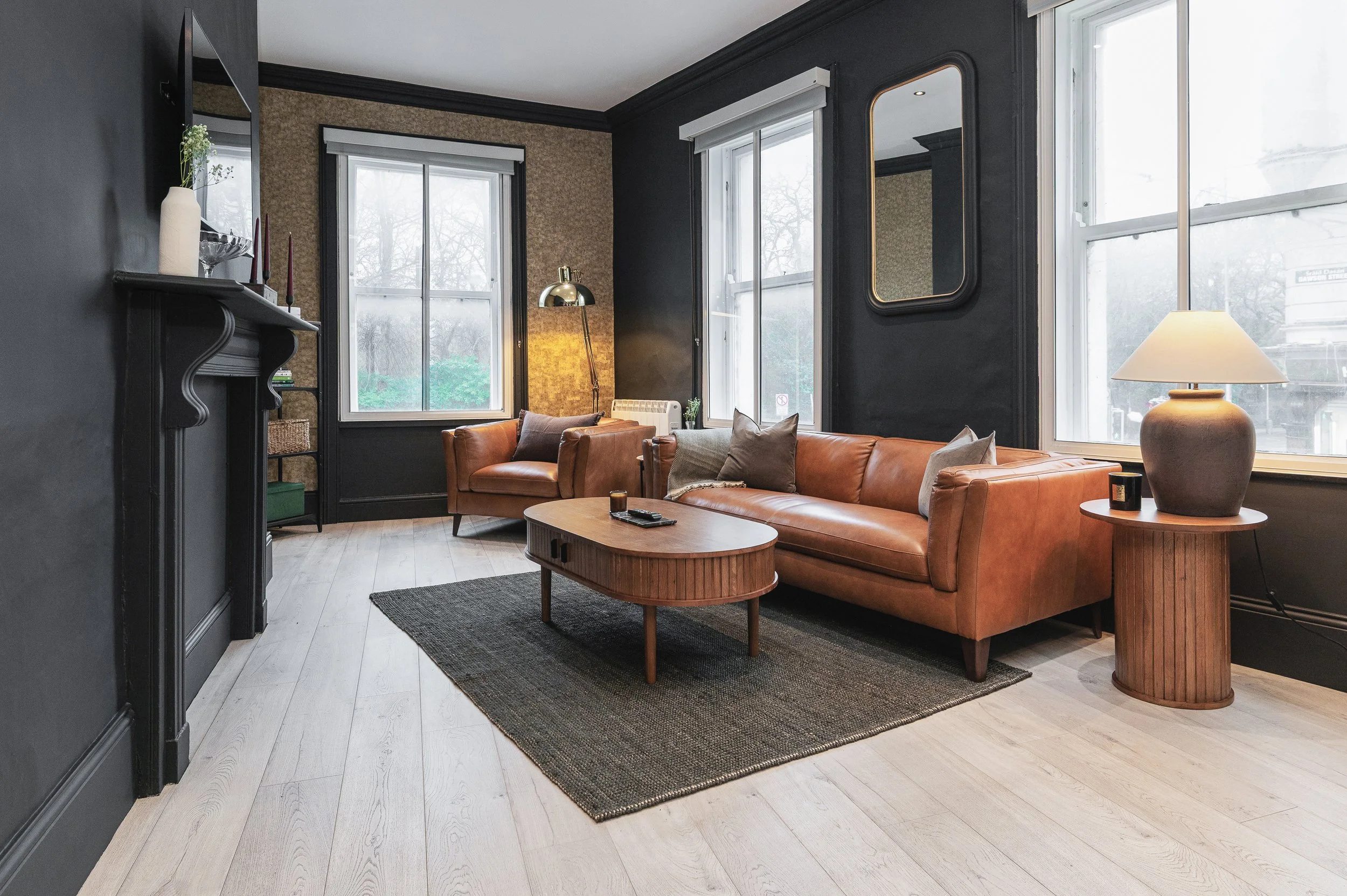 A modern living room with black accent walls and large windows. It features a tan leather sofa with gray and brown cushions, a wooden coffee table, a floor lamp with a metallic shade, and various decorative items, including a mirror on the wall.