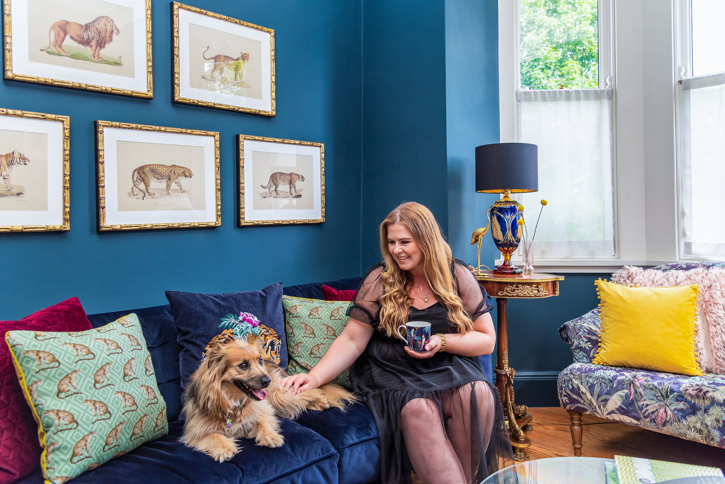 A woman sitting on a colorful couch with a gold retriever dog in a vibrant living room. The woman is smiling, holding a mug, and petting the dog. The wall behind her has five framed animal illustrations, and there is a window with green trees outside