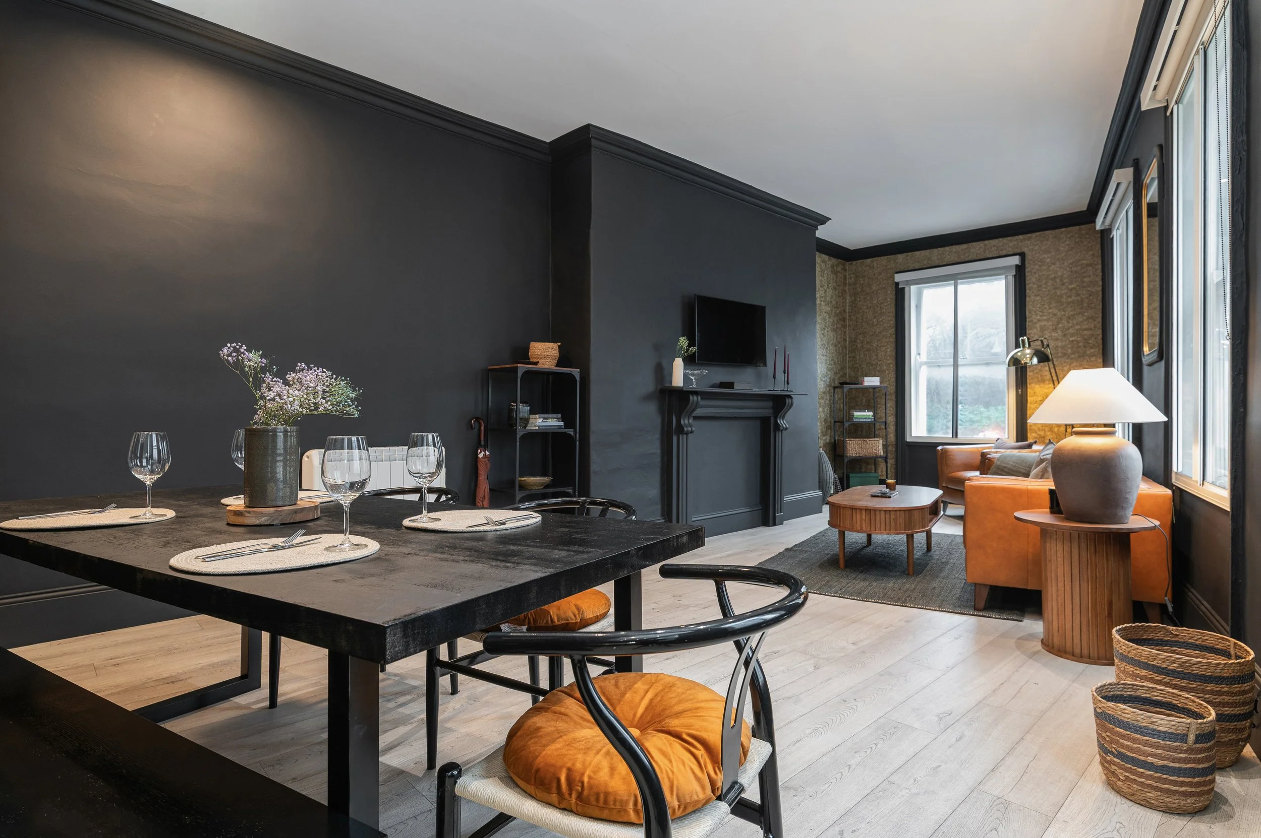 Interior of a living and dining room with dark walls, large windows, a black dining table with place settings, a floral arrangement, and a seating area with an orange sofa, a side table, and a fireplace.