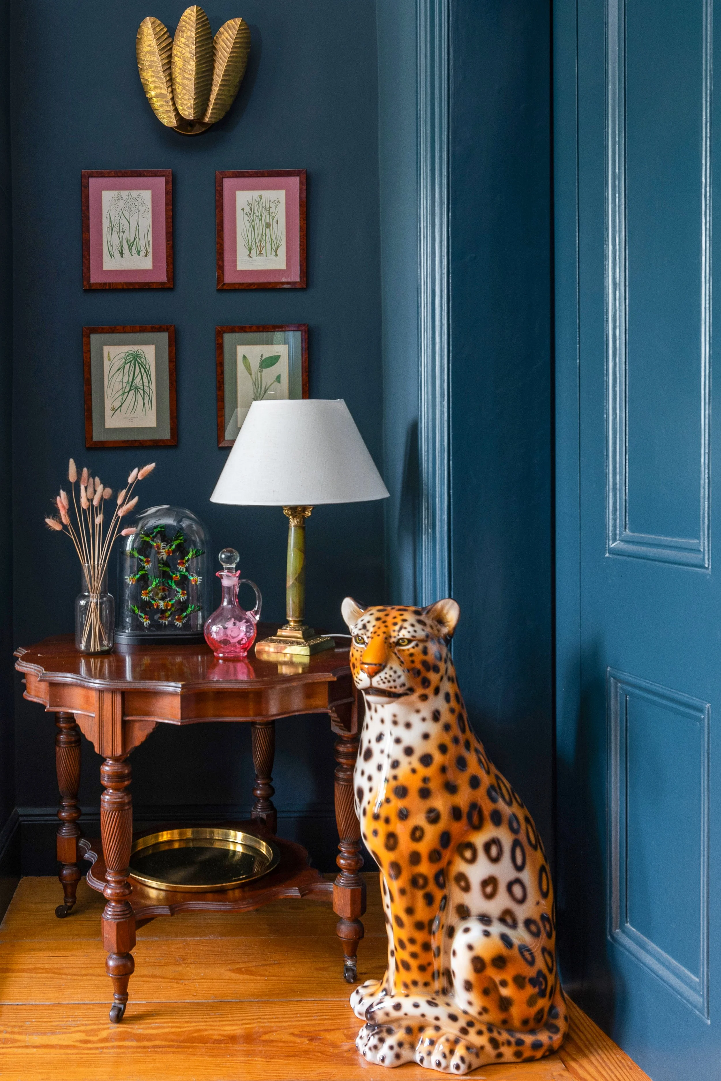 A room with dark blue walls decorated with framed botanical illustrations and a metallic gold wall art piece resembling large leaves. A wooden table holds a white lamp, decorative glassware including a pink vase and a glass dome with colorful ornamen