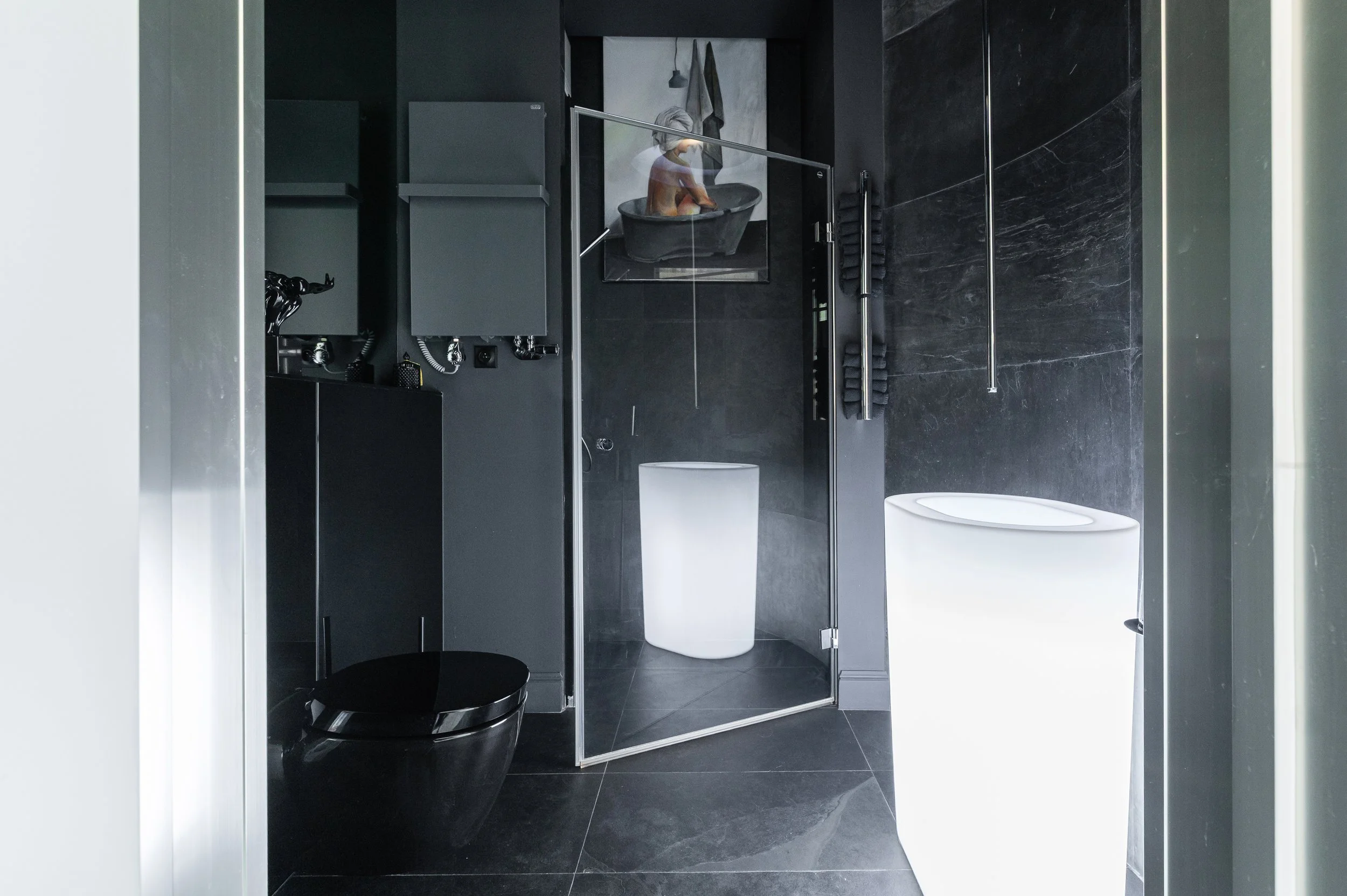Modern bathroom with black tiles, a white bidet, and a black toilet. There is a glass shower screen, a towel hanging on the wall, and artwork depicting a person in a bathtub.