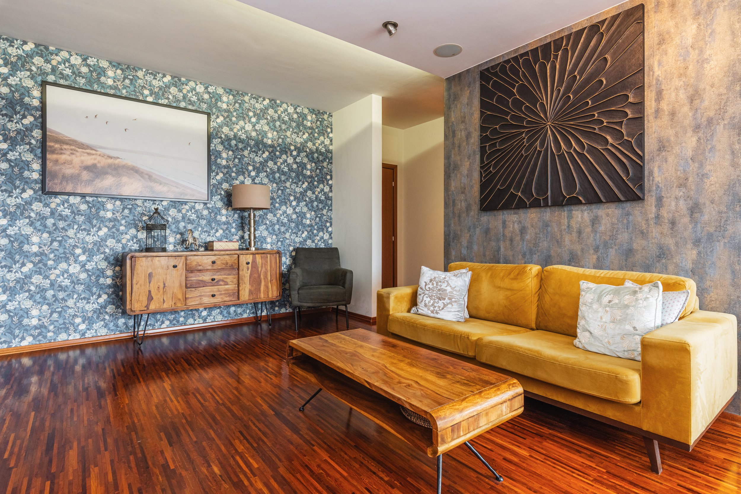 Living room with blue floral wallpaper, a wooden sideboard, a floor lamp, a gray armchair, yellow sofa with patterned cushions, large wall art, and hardwood floors.