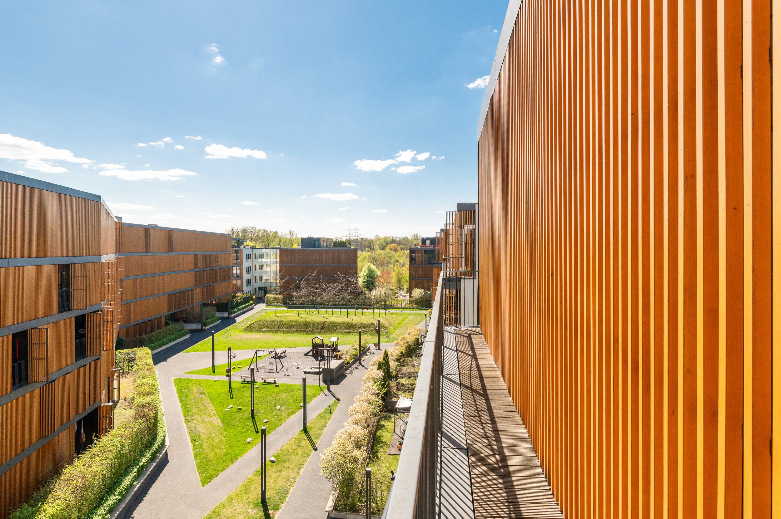 View of modern apartment complex with orange wooden siding, landscaped courtyard, walking paths, and outdoor exercise equipment under a blue sky with scattered clouds.