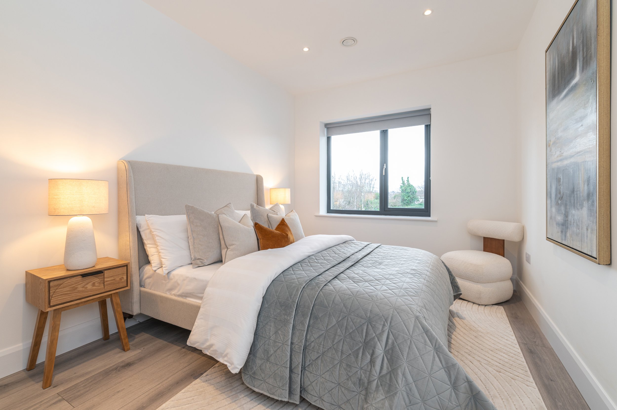 Modern bedroom with a bed, pillows, a nightstand with a lamp, a window with a view of trees, and a large abstract painting on the wall.