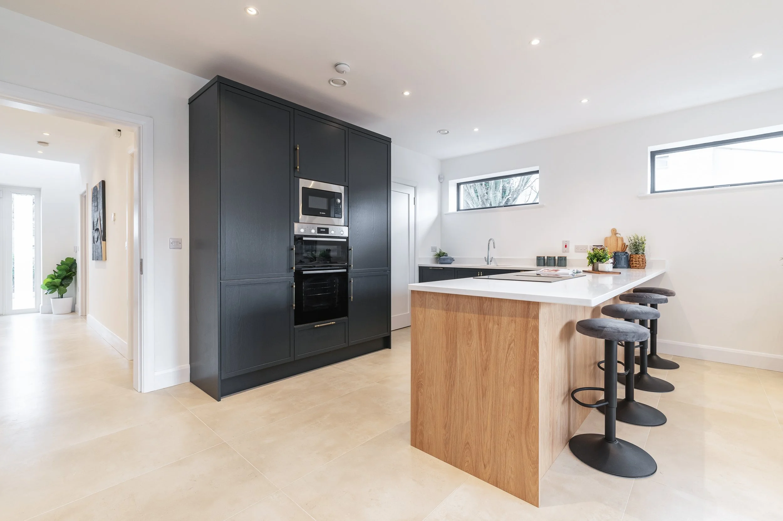 Modern kitchen with white walls, black cabinetry, a white countertop island with wooden sides, four black bar stools, and windows with trees outside.