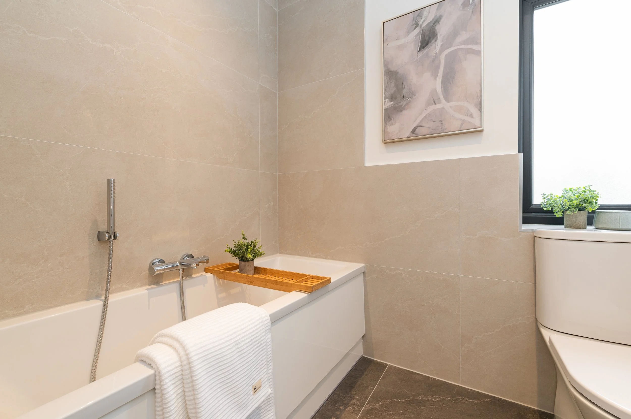 Modern bathroom with beige marble tiles on walls and dark gray tiles on floor. Features a bathtub with a white towel draped over the edge, a small wooden tray, potted plants, a large abstract painting on the wall, a window with a green plant on the w
