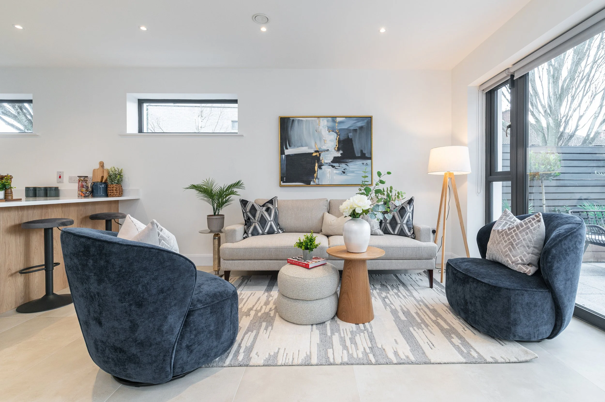 Modern living room with a sofa, armchair, coffee table, plants, artwork, and a floor lamp near large glass windows.