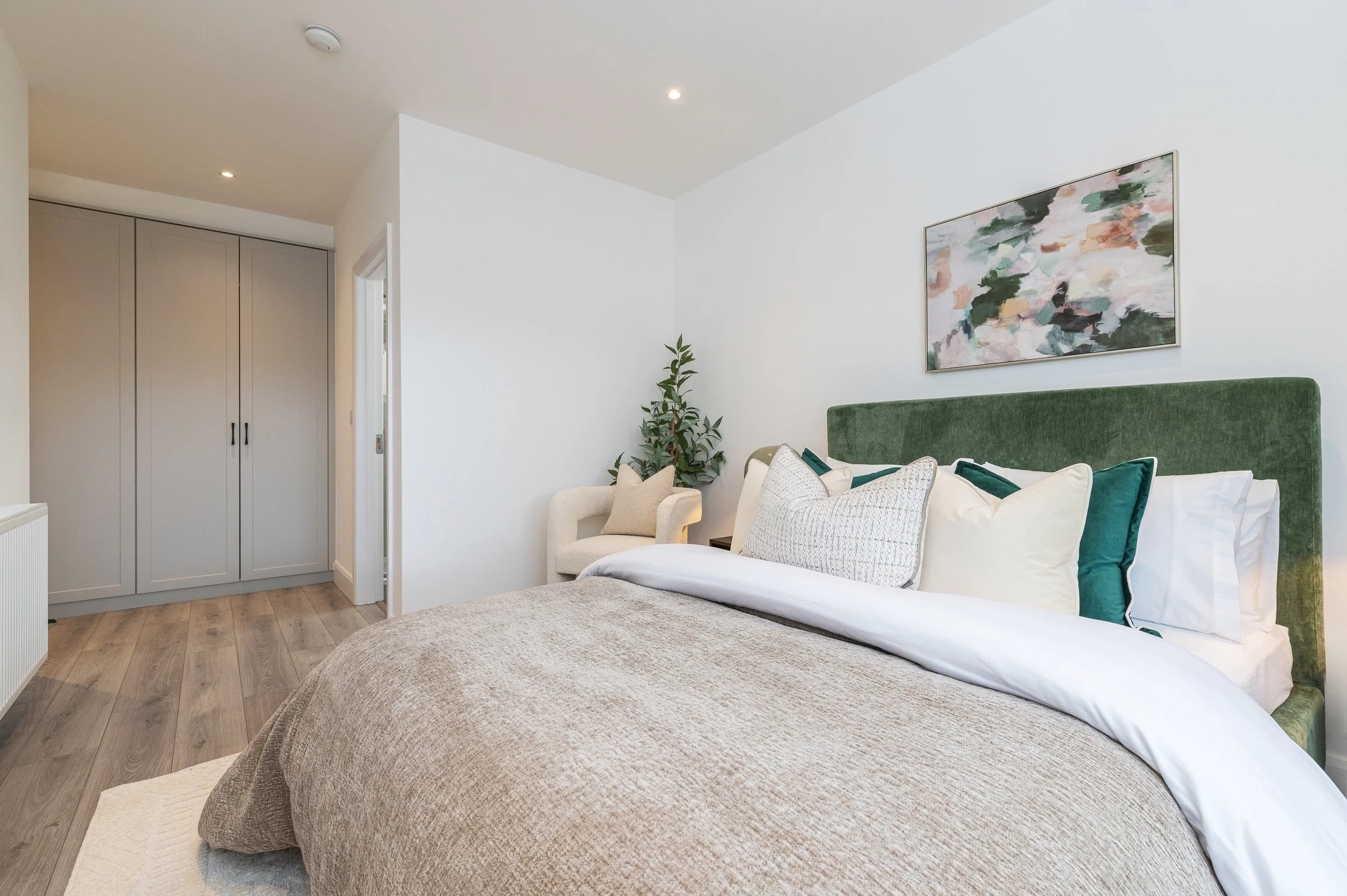 A modern bedroom featuring a bed with a green velvet headboard, white and beige pillows, and a beige blanket. A wall-mounted abstract painting hangs above the bed. There is a small beige armchair with pillows, a potted plant next to it, and a built-i