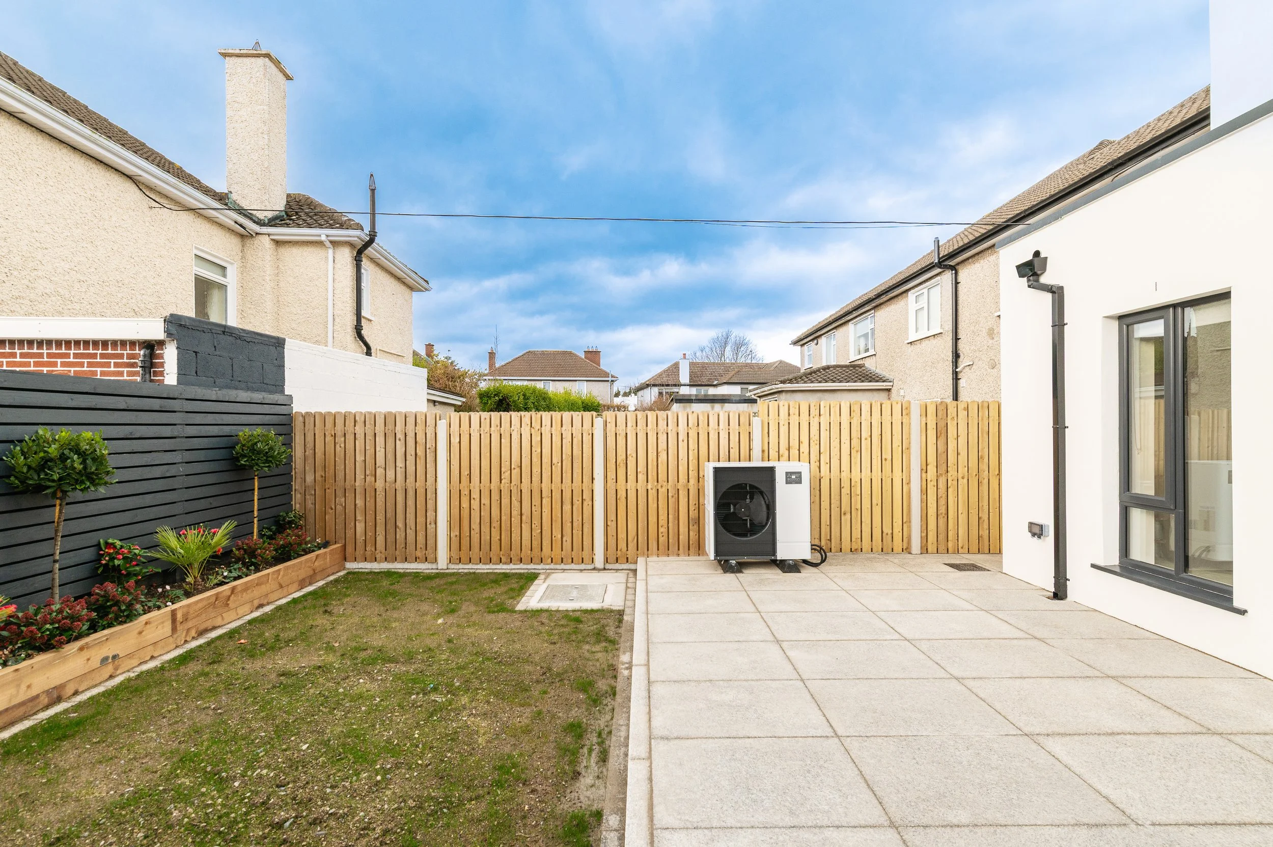 A backyard with a small garden bed, a fence, and neighboring houses under a partly cloudy sky.