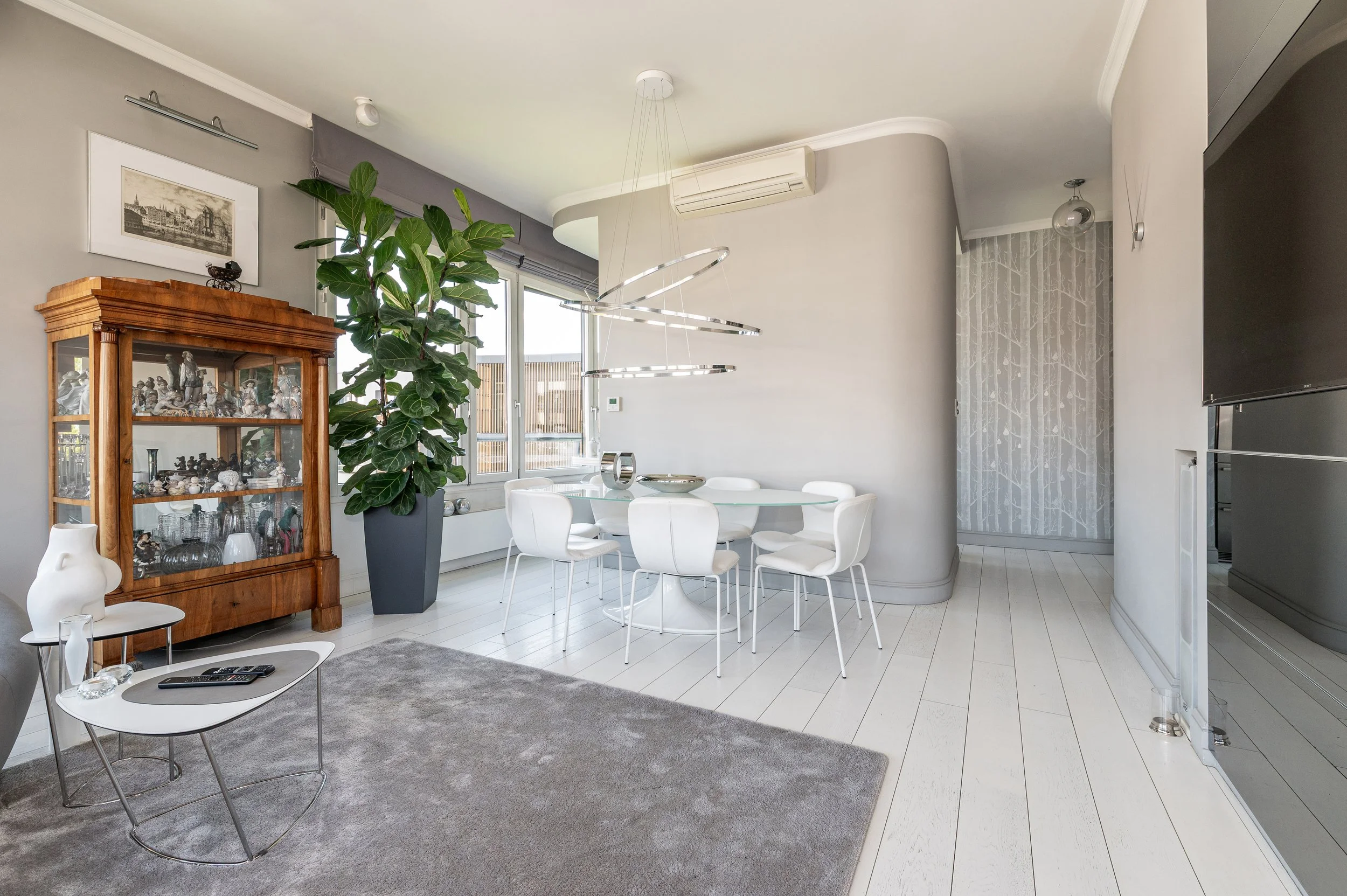 Modern living and dining area with white circular dining table and chairs, gray area rug, large green plant, wooden display cabinet with figurines, large window, and contemporary hanging light fixture.