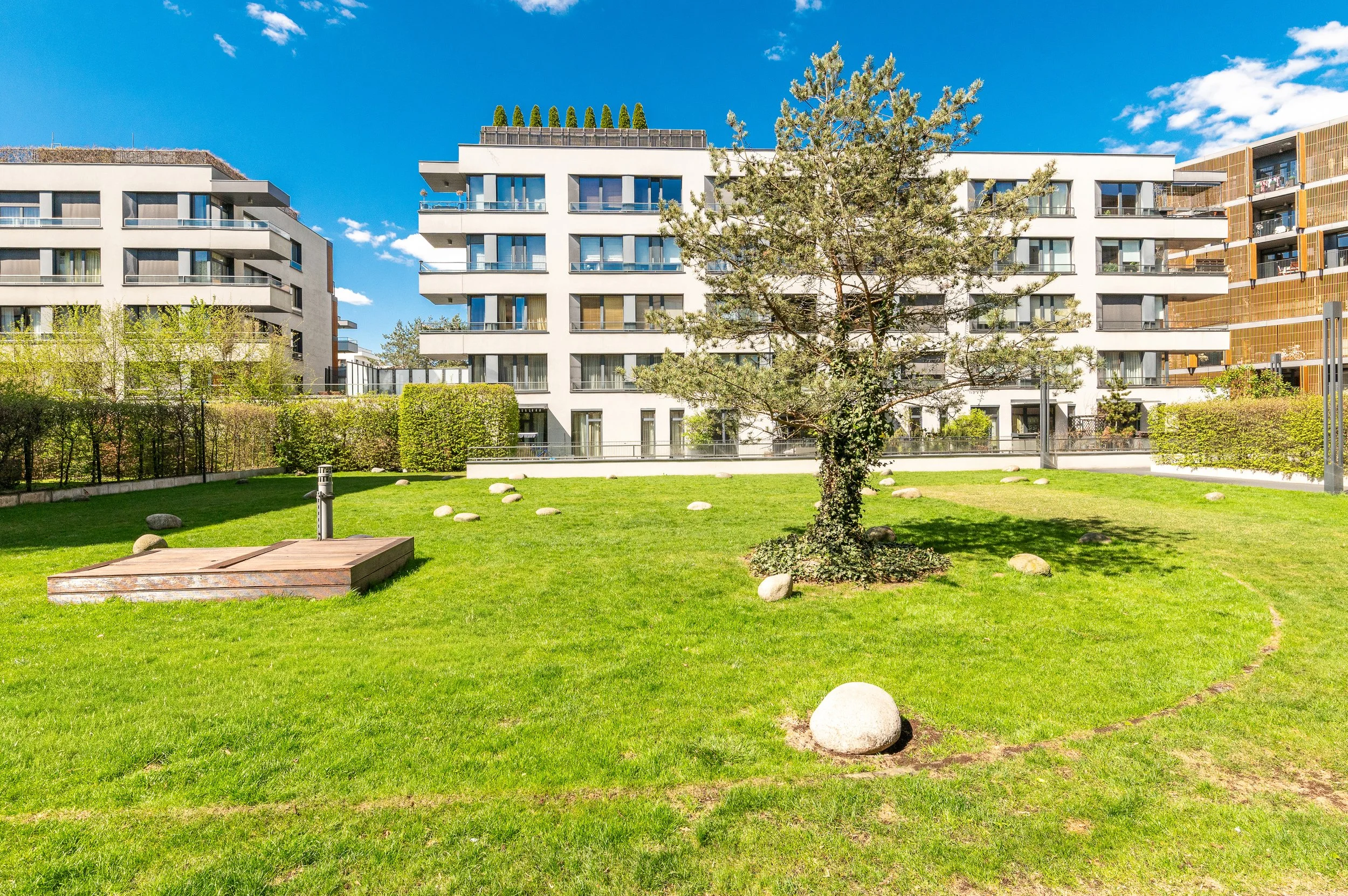 Modern white apartment buildings with balconies and large windows, green lawn with rocks and a small tree in the foreground, blue sky with a few clouds.