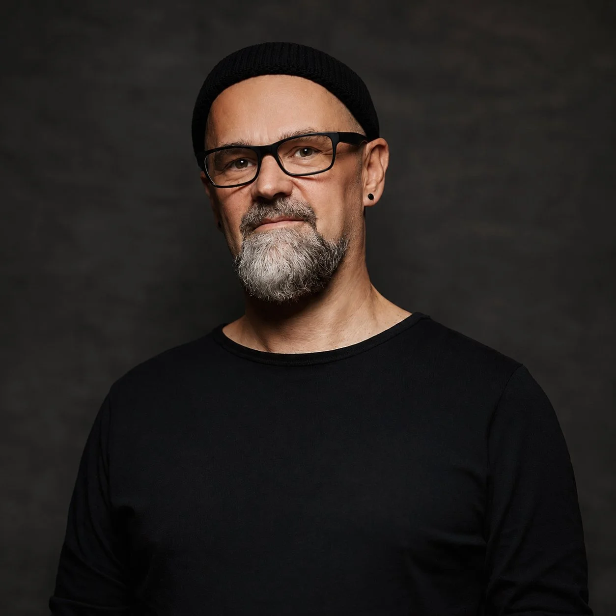 Portrait of a middle-aged man with glasses, a beard, and a mustache, wearing a black beanie and black shirt, against a dark background.