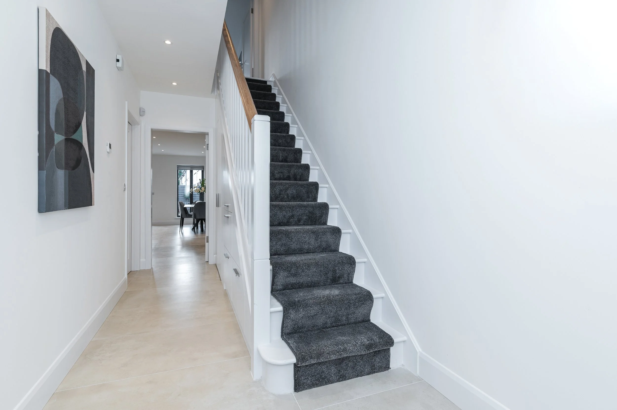Interior of a modern home with a staircase featuring dark carpeted steps, white risers, and a wooden handrail. The hallway has white walls, recessed lighting, and light-colored tiled flooring. A piece of abstract artwork hangs on the left wall, and a