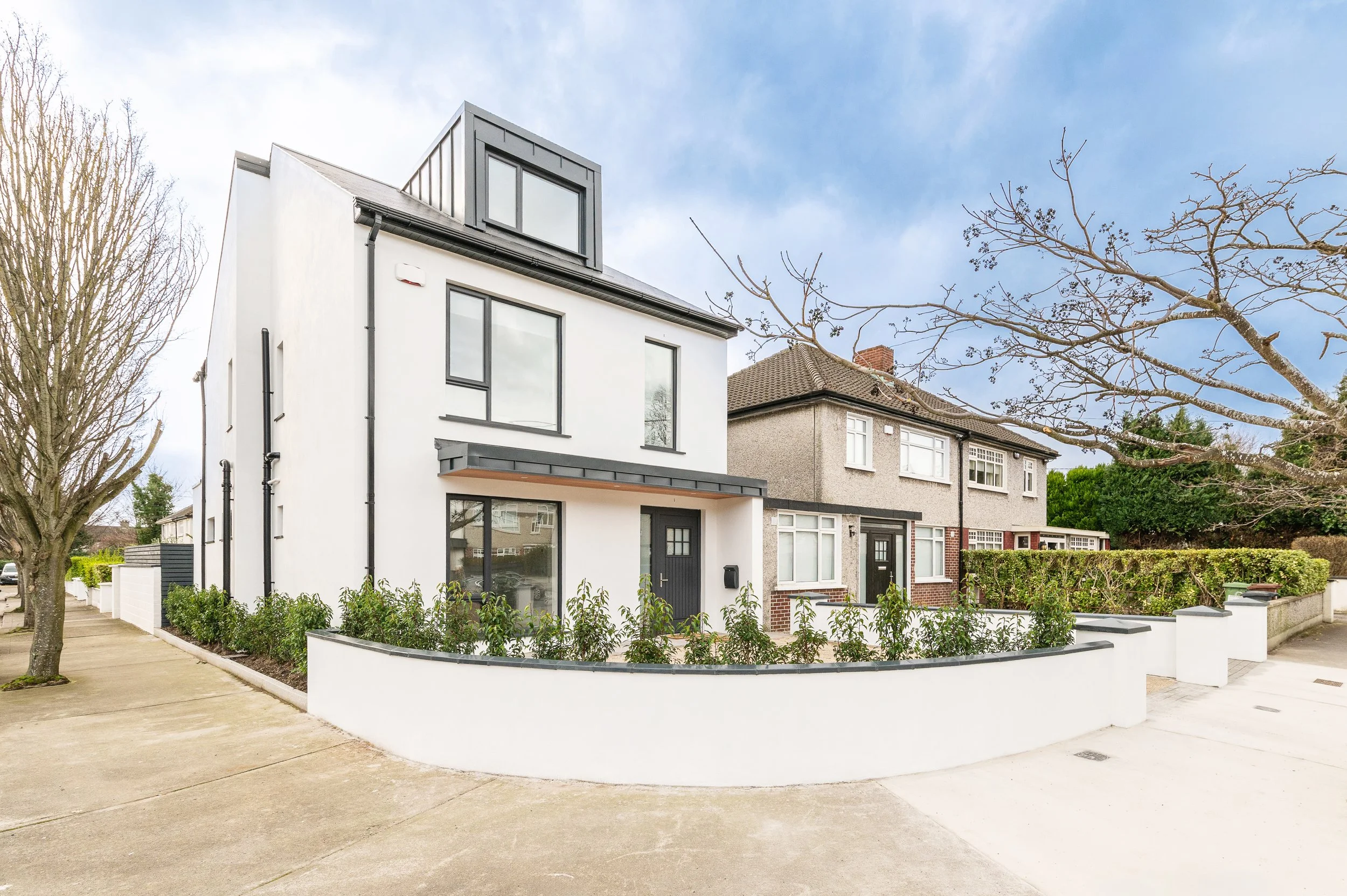 Modern white house with black window frames, front garden with bushes, surrounding sidewalk, leafless trees, neighboring houses, overcast sky.
