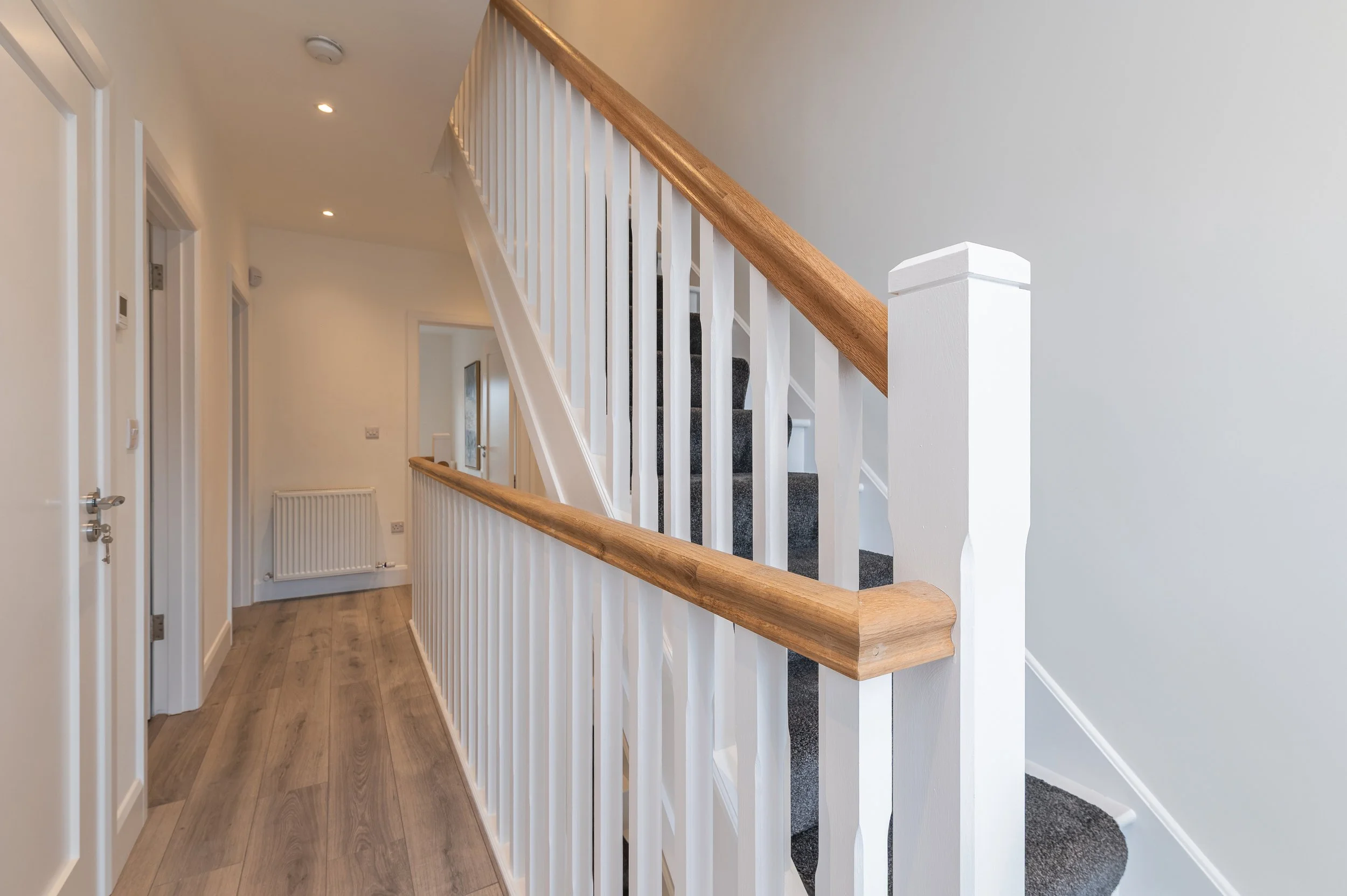 Interior view of a hallway with a staircase featuring white balusters and a wooden handrail, with hardwood floors, white walls, and ceiling lights.