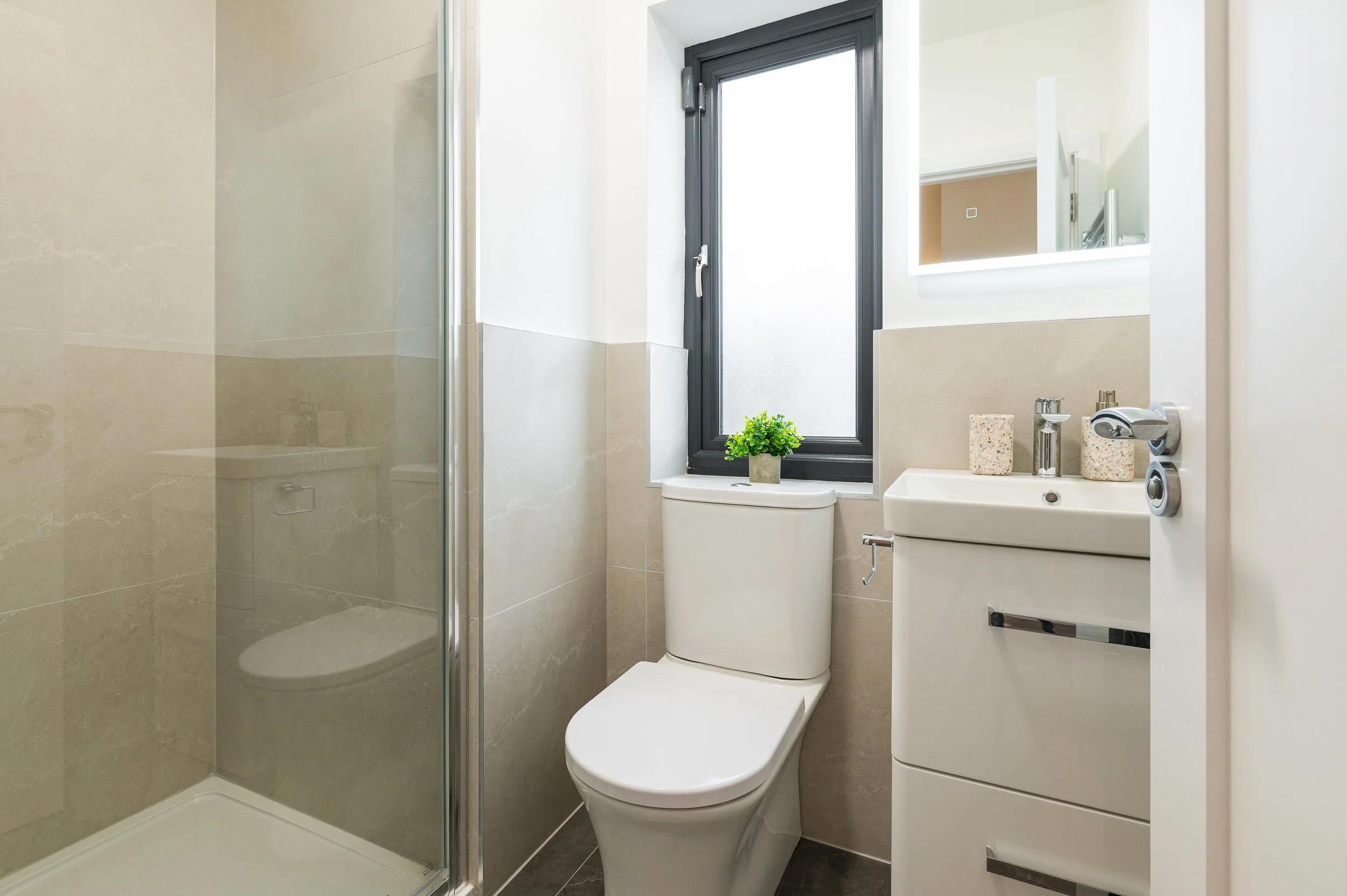 Modern bathroom with a shower stall, a toilet, a small vanity with a sink, a mirror with built-in lighting, a window with a small potted plant on the toilet tank, and beige tiled walls.