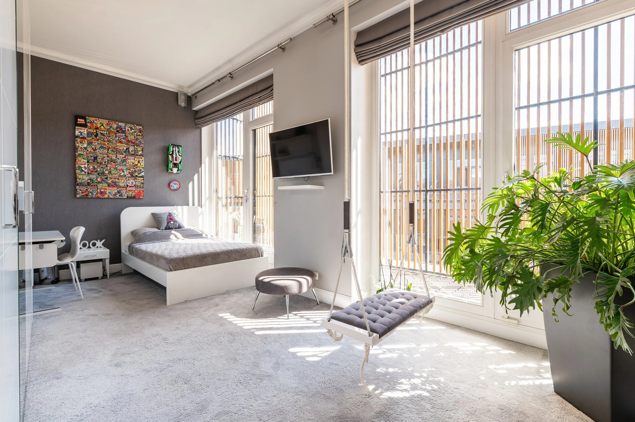 Bright bedroom with large windows, sliding glass door, and gray curtains. Contains a bed with gray bedding, a white desk, a swivel chair, a wall art showcasing comic books, a small wall-mounted TV, a gray ottoman swing, and a large green leafy plant.