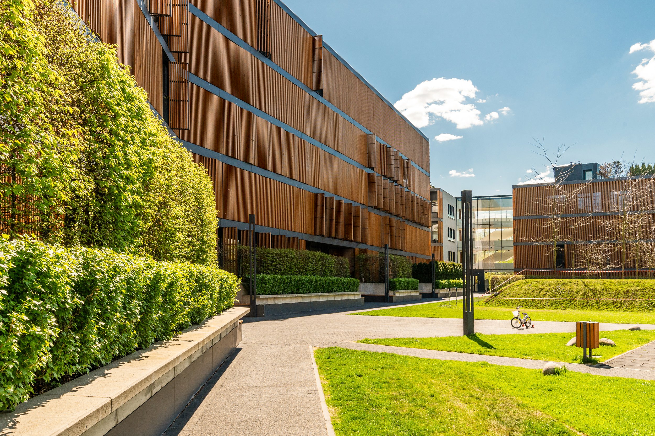 Modern multi-story building with wooden exterior panels, surrounded by a well-kept park with green grass, trees, bushes, a sidewalk, a bicycle, and a clear blue sky with some clouds.