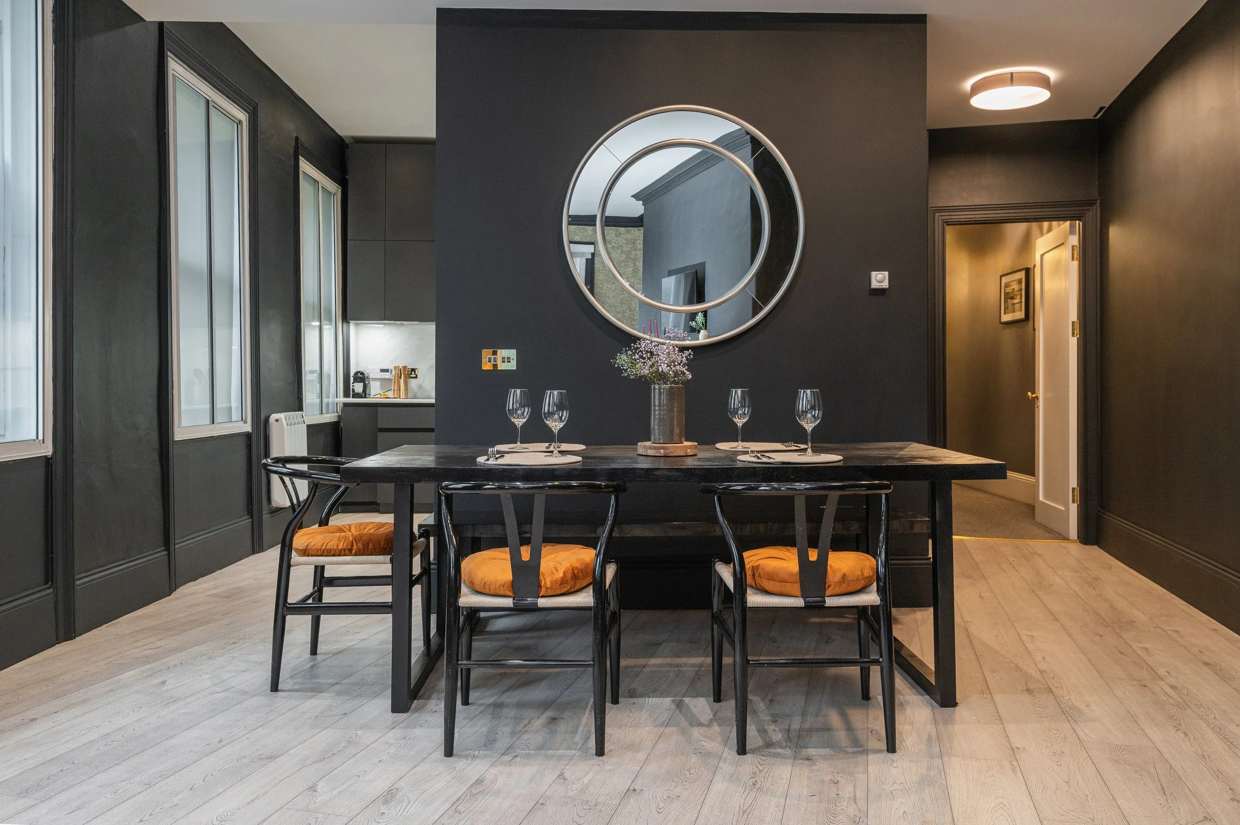 A dining room with a dark wooden table set for four, with wine glasses and placemats. Four chairs with orange cushions surround the table. A large round mirror hangs on the dark-colored wall behind the table, reflecting the outdoors. To the left, par