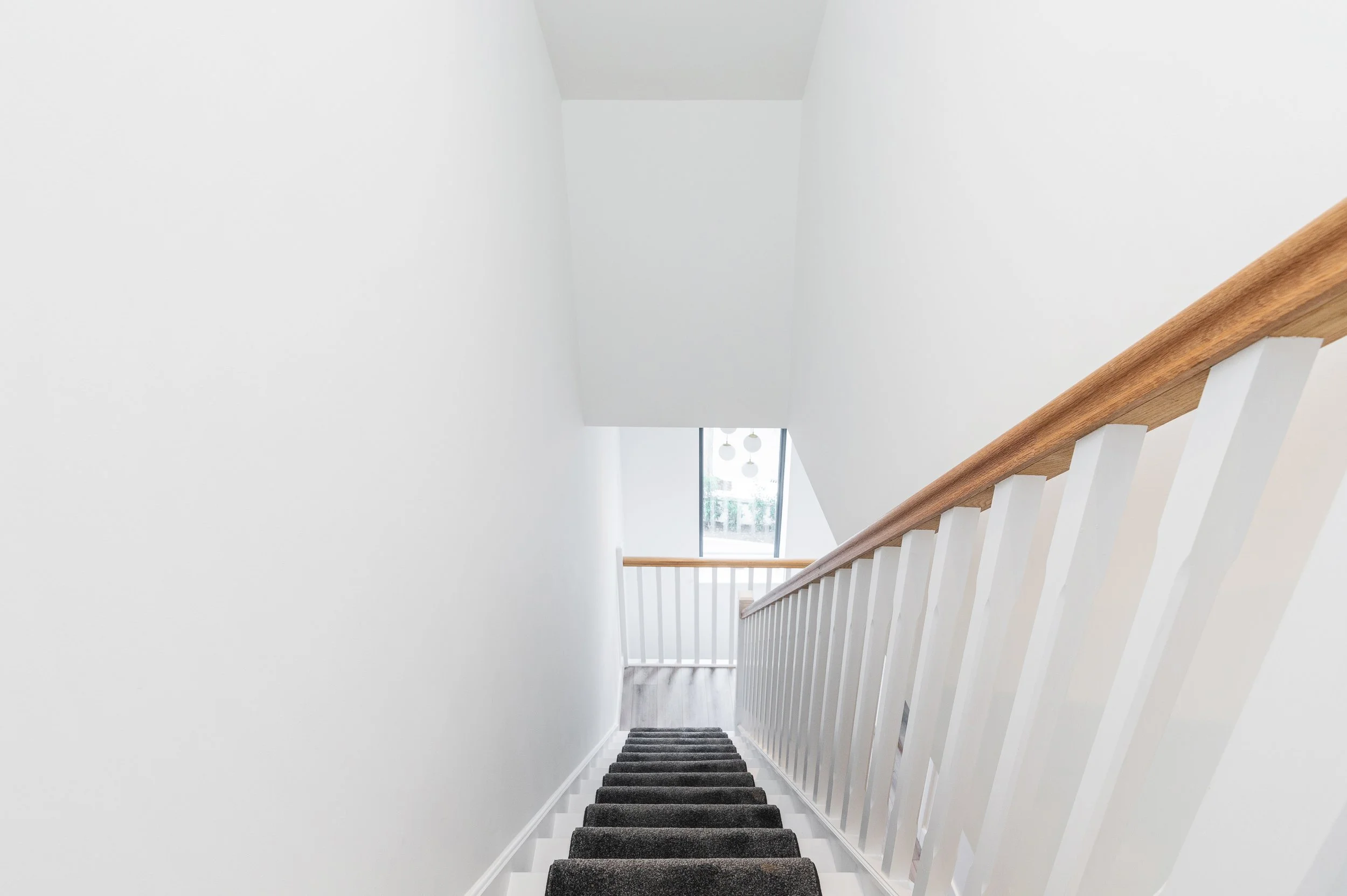 Interior view of a staircase with gray carpeted steps, white walls, a wooden handrail on the right, and a window with hanging light fixtures at the top of the stairs.