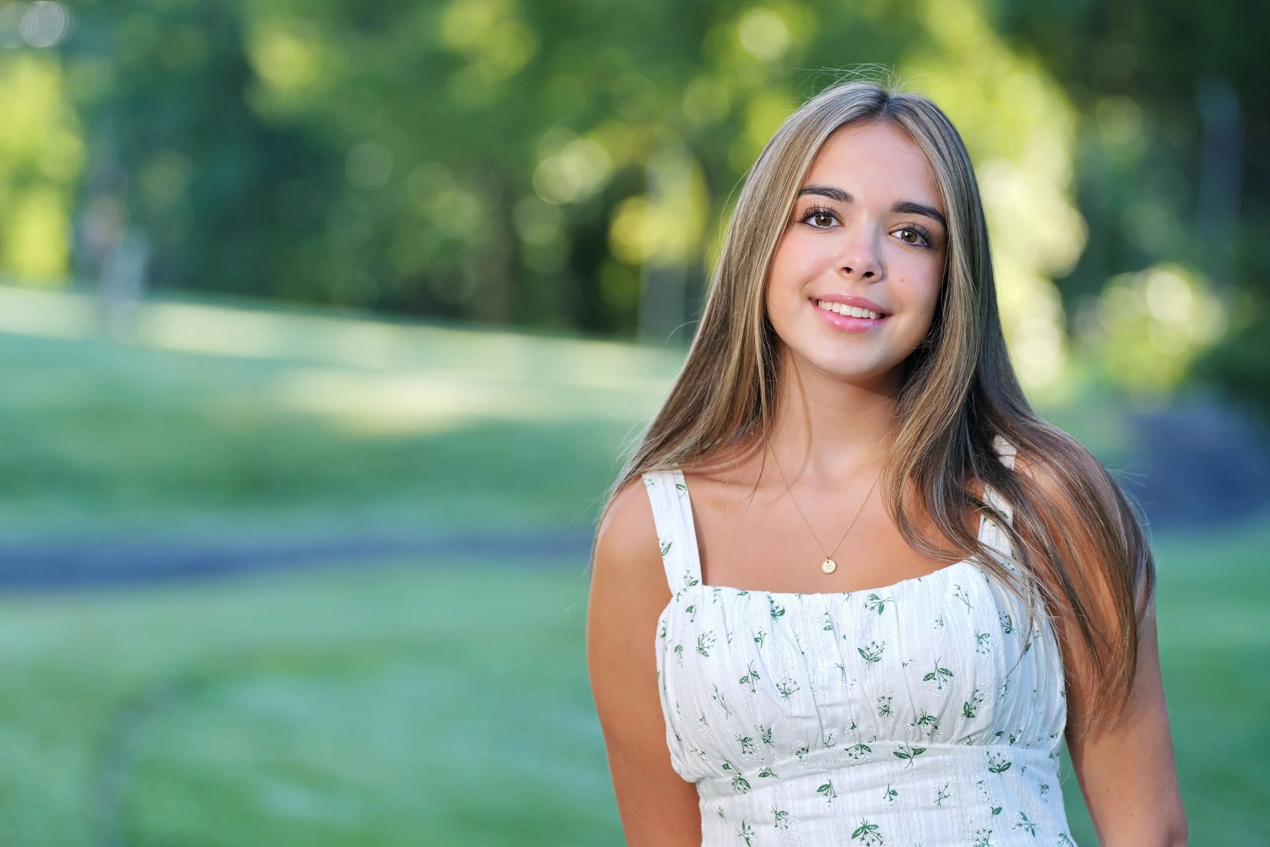 A young woman with long brown hair and brown eyes smiling outdoors in a green park during daylight.