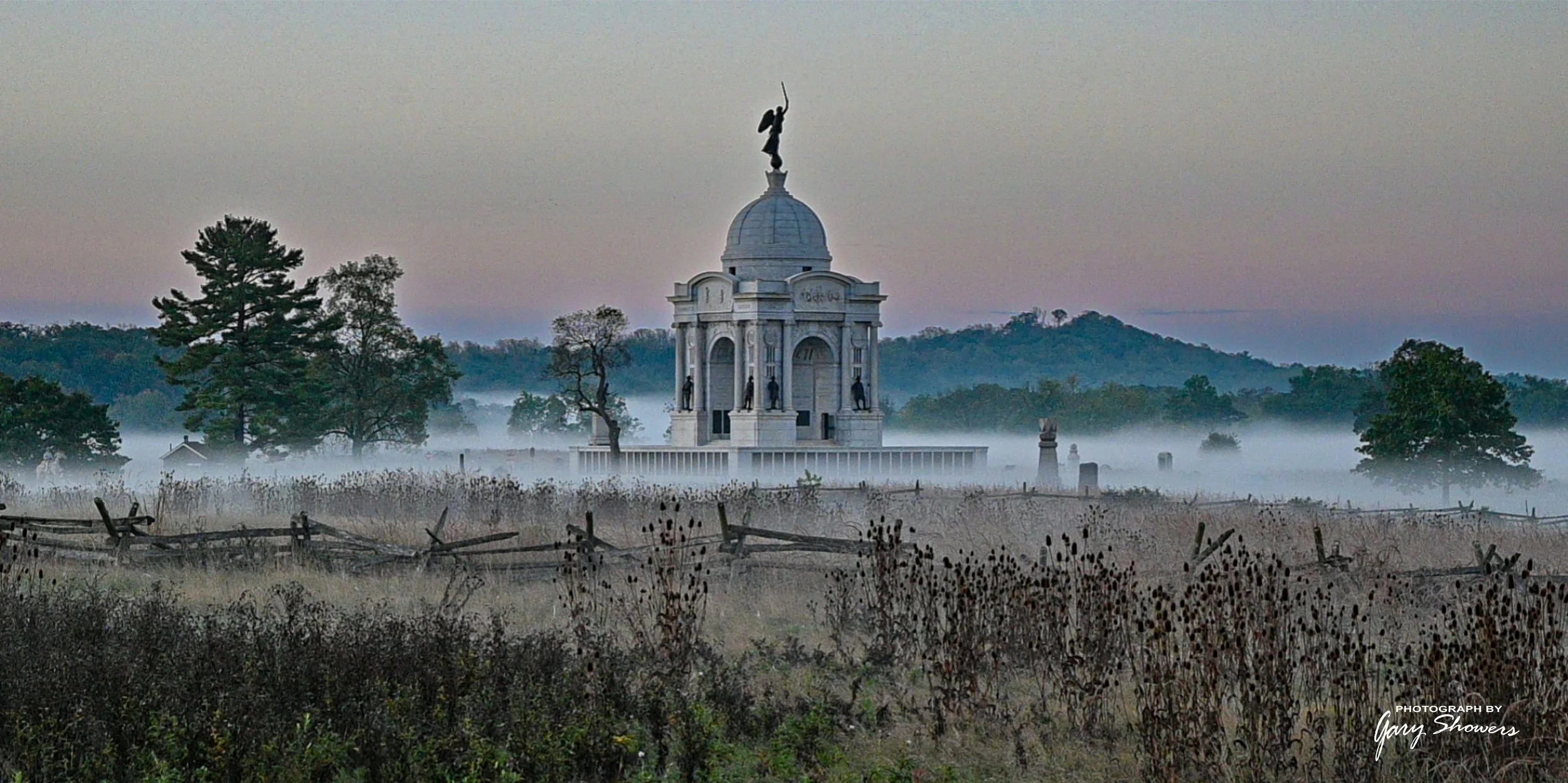 A tall, white monument with statues and a dome features a statue on top, set in a misty rural landscape with trees, hills, and a foggy sky at dawn or dusk.