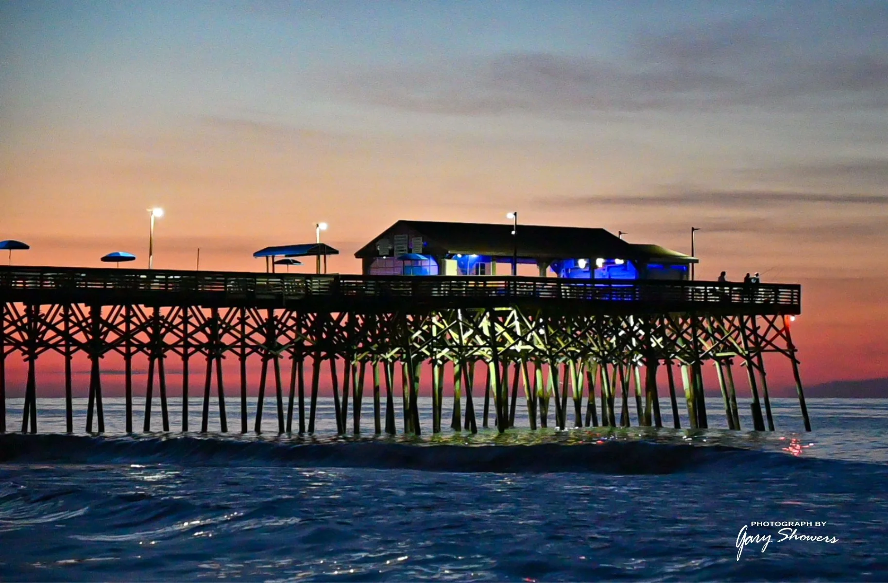A pier extends into the ocean at sunset with colorful sky in the background. The pier has a small building with blue lighting and several umbrellas, with a few people visible on the railings.