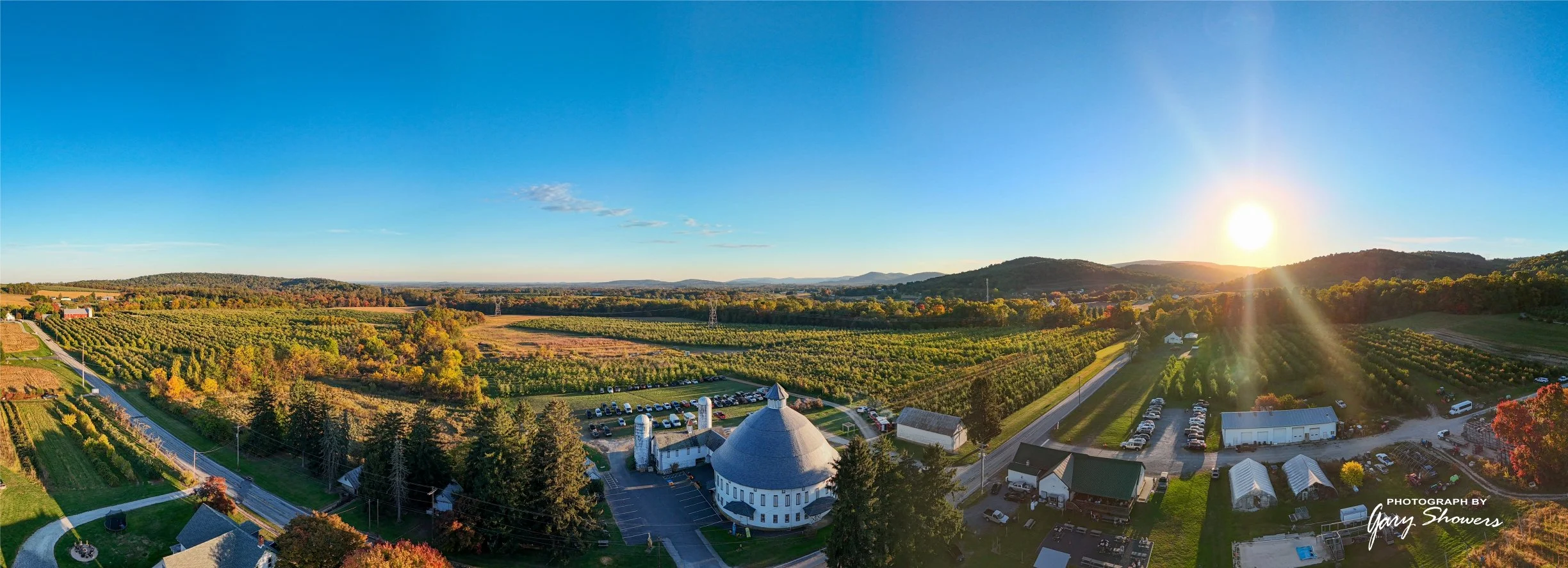 A scenic aerial view of farmland during sunset with vineyards, trees, buildings, and a clear sky.