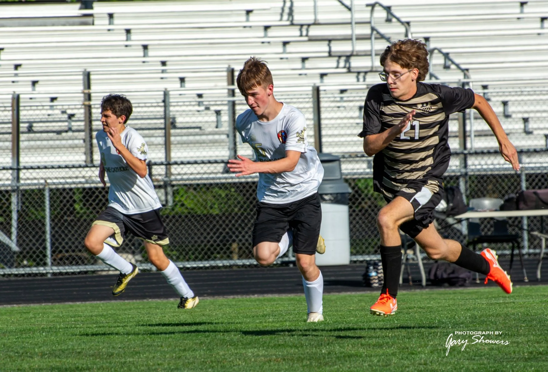 Three young boys running on a soccer field during a game or practice, with empty bleachers in the background.