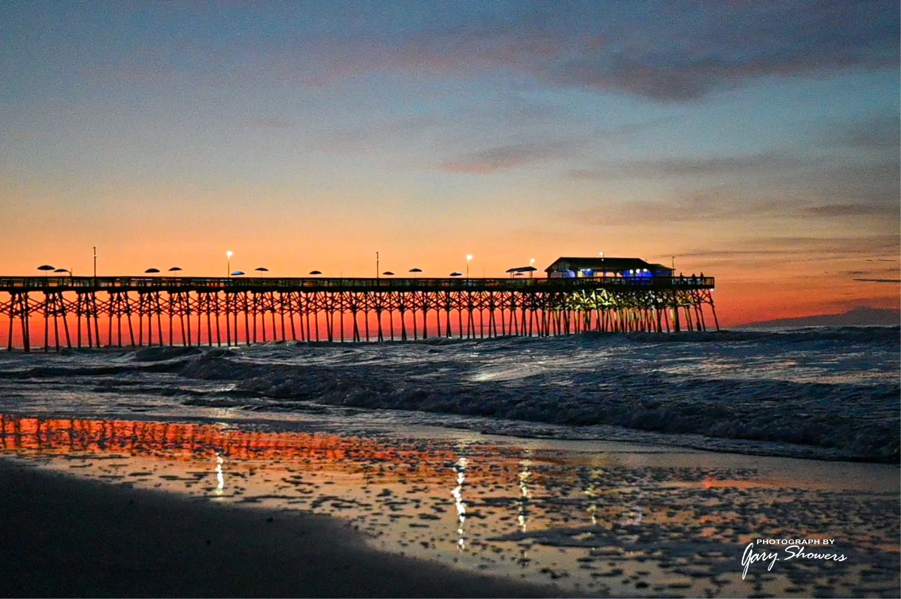 A pier extending into the ocean during sunset, with umbrellas on the pier and lights illuminating a building at the end of the pier, reflected on the wet sand and water below.
