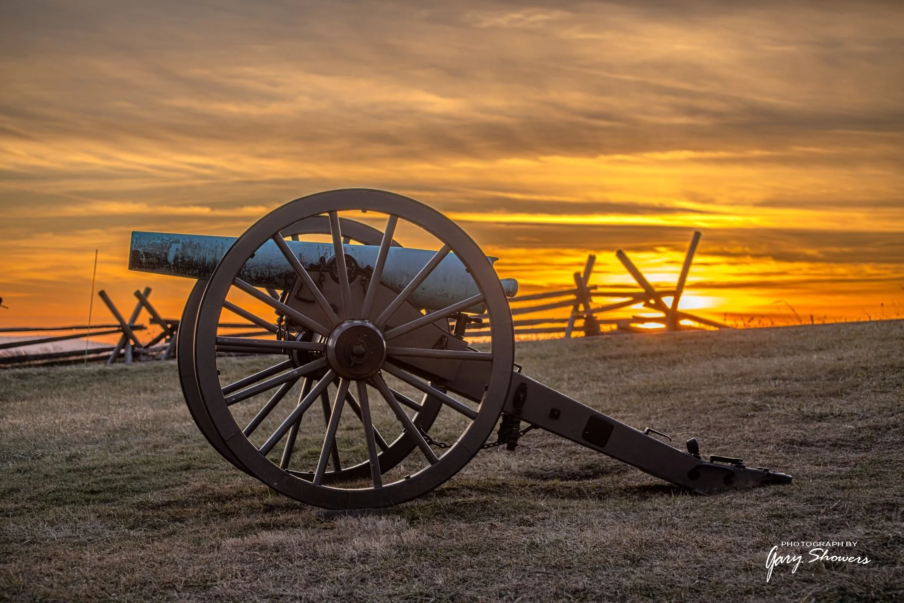 An old cannon on a grassy hill at sunset with a wooden fence in the background.