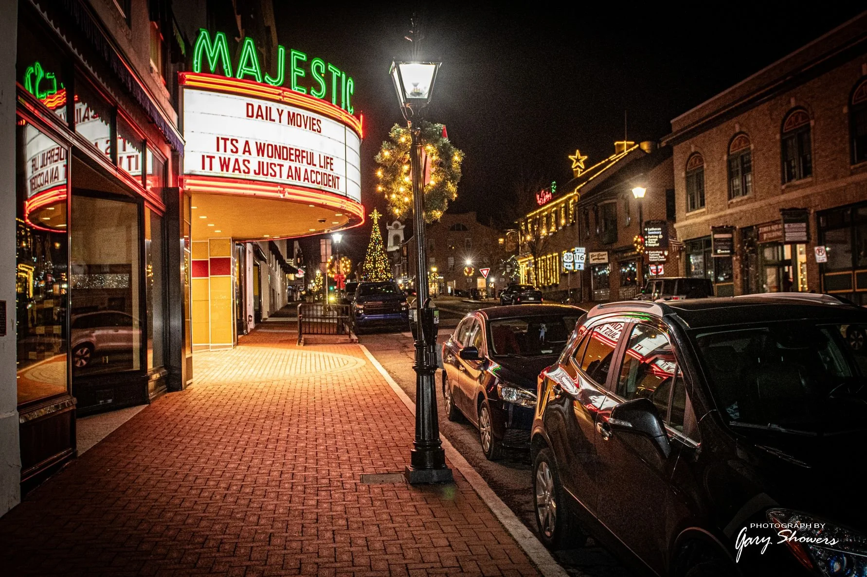 Empty sidewalk and street at night in a small town decorated with Christmas lights. A marquee sign displays movie titles, and holiday decorations including a lit Christmas tree and wreaths are visible.