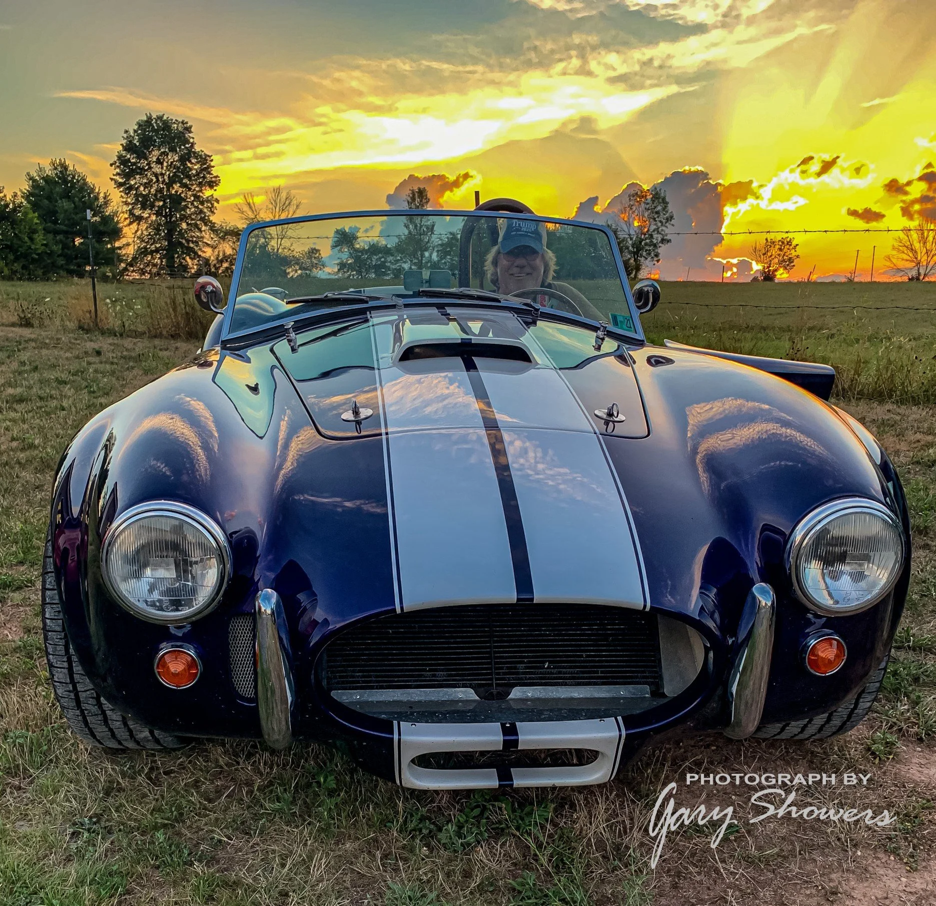 A classic black sports car with white racing stripes parked on grass during sunset, with a person wearing a cap sitting inside, reflected in the windshield.