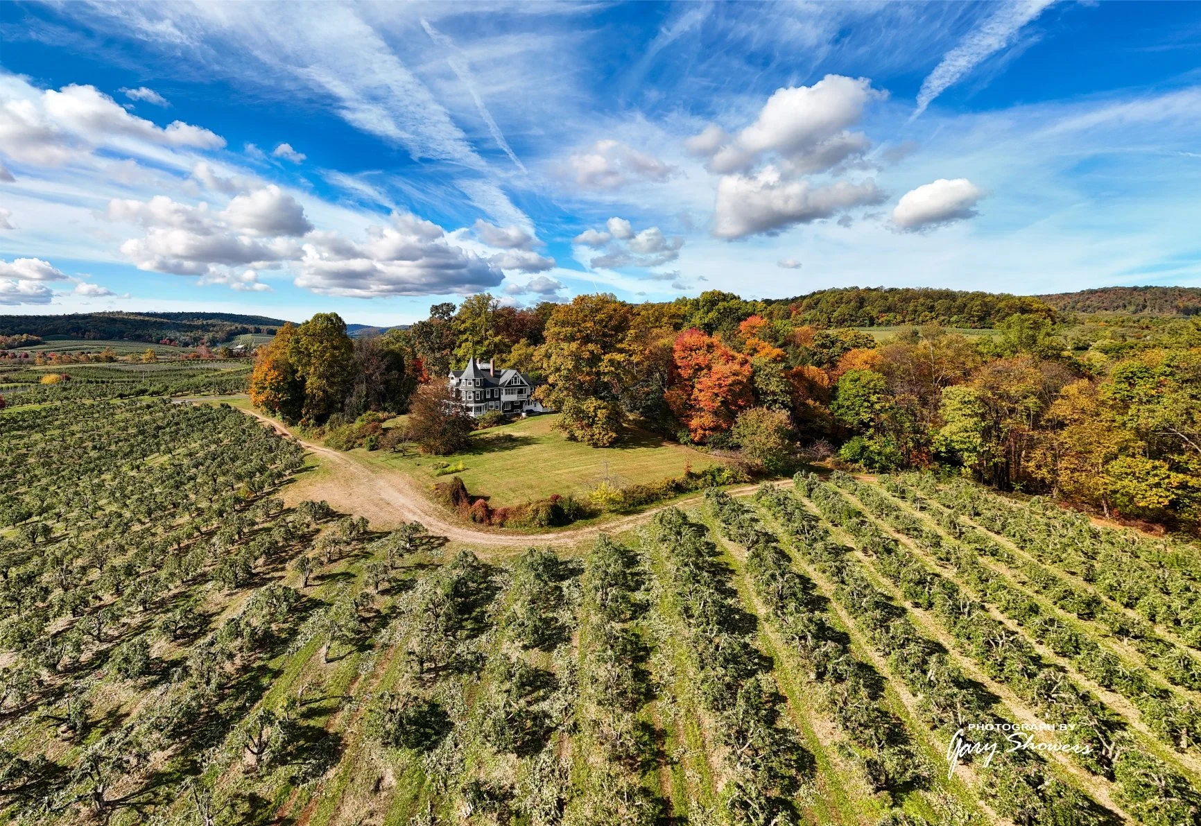 Landscape view of a large house surrounded by colorful fall trees and a vineyard under a partly cloudy sky.