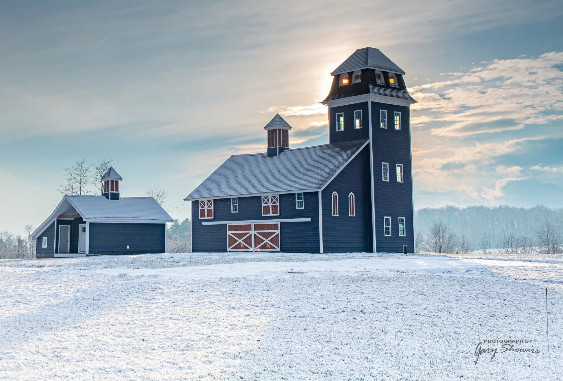 A large black barn with a tower, emitting light from some windows, and a smaller black outbuilding, both surrounded by snow on the ground, under a cloudy sky during sunset.