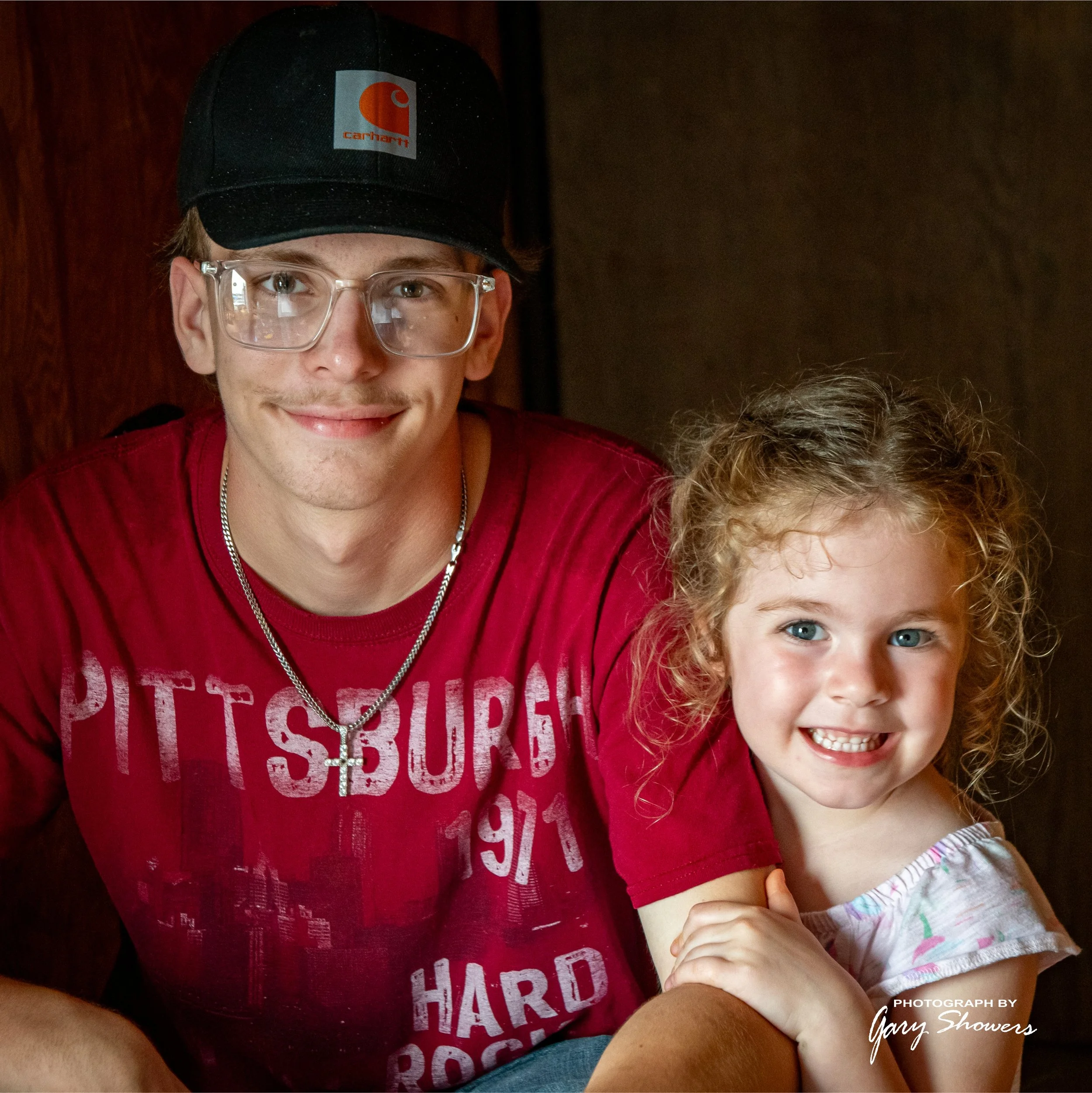 A young man with glasses, wearing a black Carhartt cap and red Pittsburg T-shirt, smiling with a young girl with curly blonde hair and blue eyes, wearing a white top with colorful patterns, both sitting closely together and smiling.
