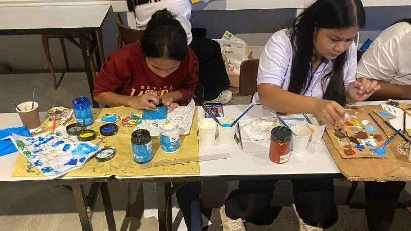 Two girls painting at a table with various art supplies, including paint bottles, brushes, and paper during an art activity.