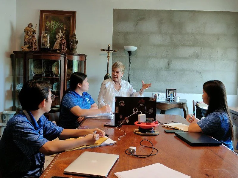 Four women seated around a wooden table in a meeting room, engaged in a discussion with a woman standing, gesturing with her hand. The table has laptops, notebooks, and chargers, with a tall cabinet and decorative statues in the background.