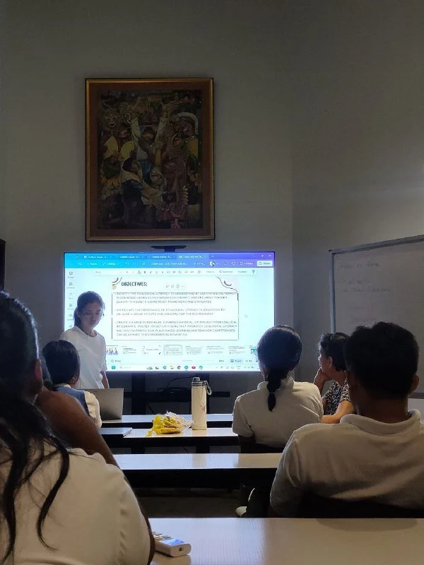Classroom with students watching a girl giving a presentation at the whiteboard, a painting hanging on the wall behind her.