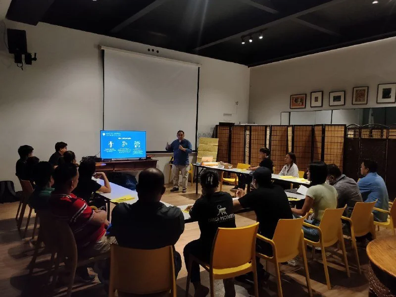 Person giving a presentation to a seated audience in a room with a large screen, artwork on the walls, and folding screens in the background.
