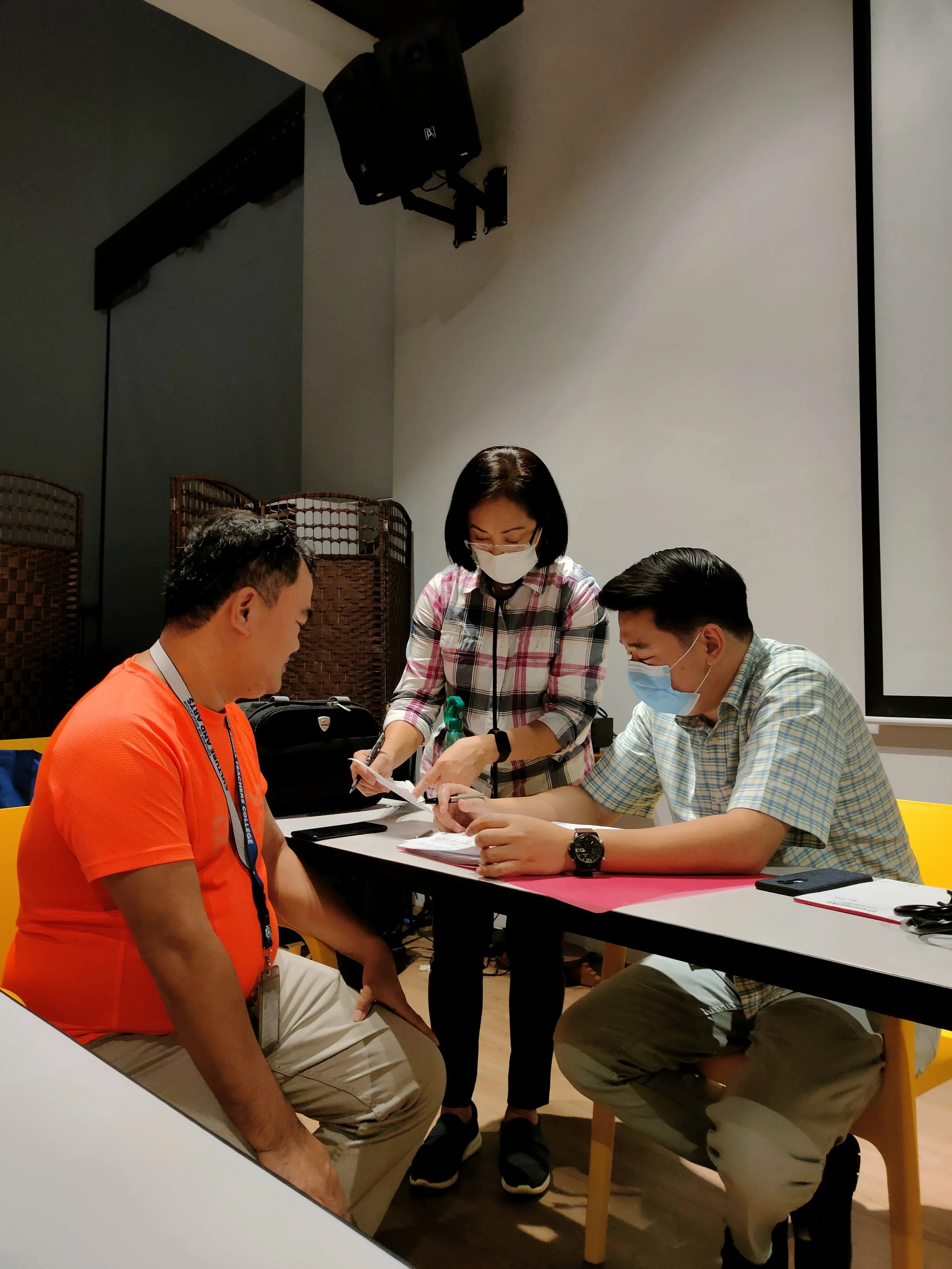 Two men sitting at a table and a woman standing between them, all wearing face masks, engaging in a discussion with documents, in an indoor setting with a large screen and a speaker in the background.