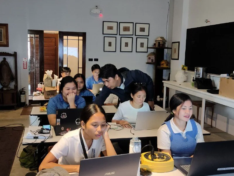 Group of students working on laptops in a classroom with a teacher assisting, students wearing blue and white uniforms, shelves and framed pictures on the walls, and a large TV screen.