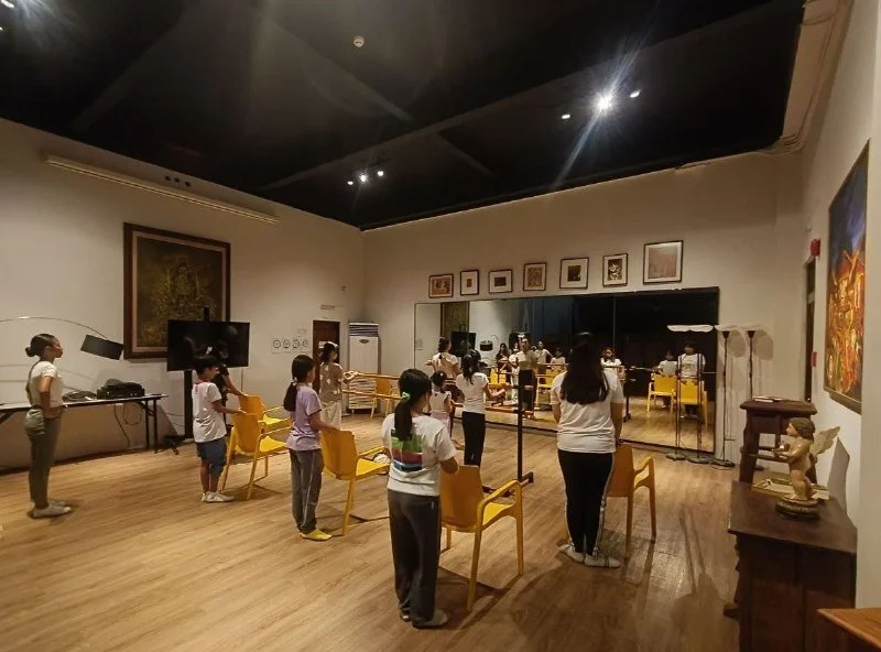 Children in face masks participating in an indoor educational activity or tour, standing behind yellow chairs, with a teacher or guide instructing them, in a room with artwork on the walls and a large mirror.