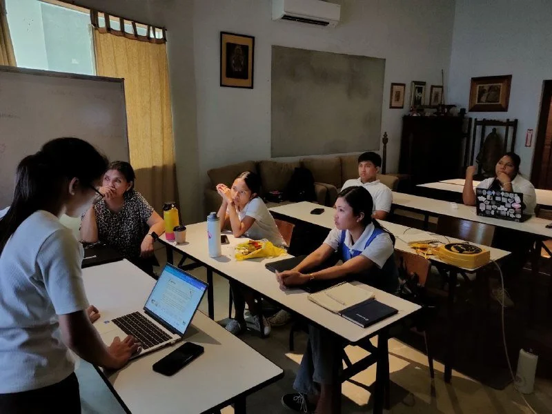Group of students attending a classroom lecture, seated at desks with laptops and notebooks, listening to a woman standing at the front.