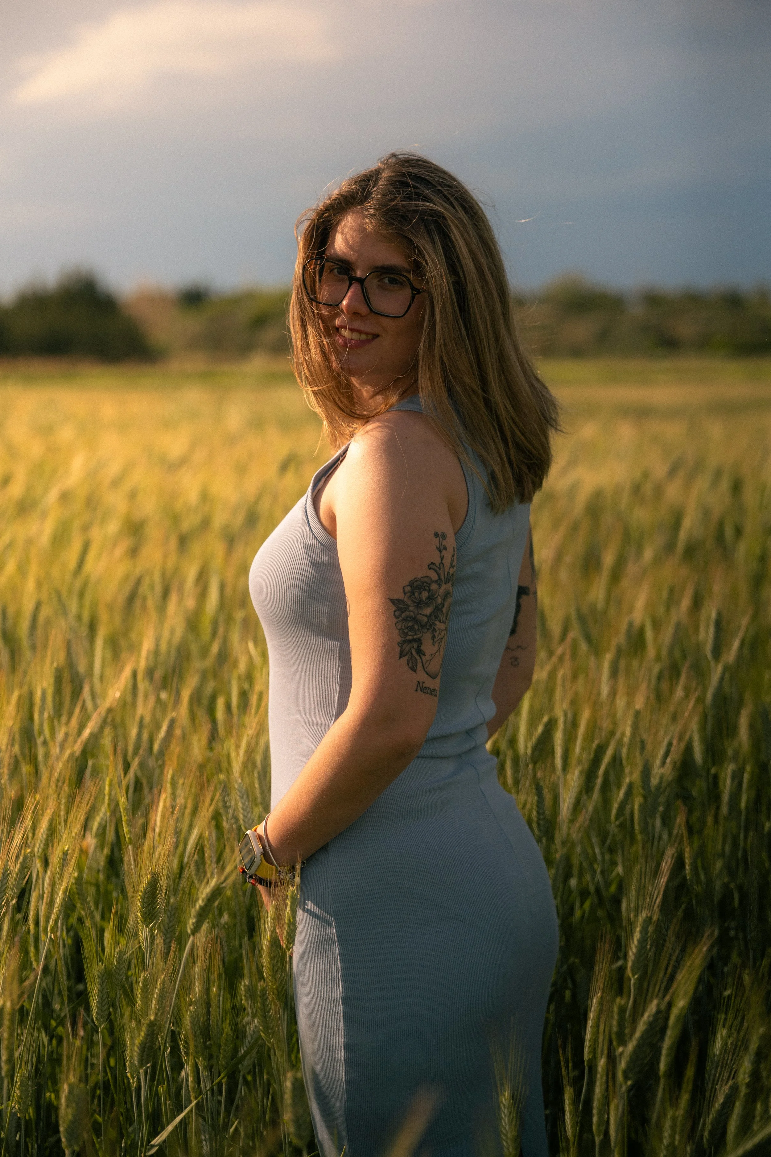 Mujer con gafas y tatuajes en el brazo, de pie en un campo de trigo durante el atardecer, sonriendo y mirando hacia la cámara.
