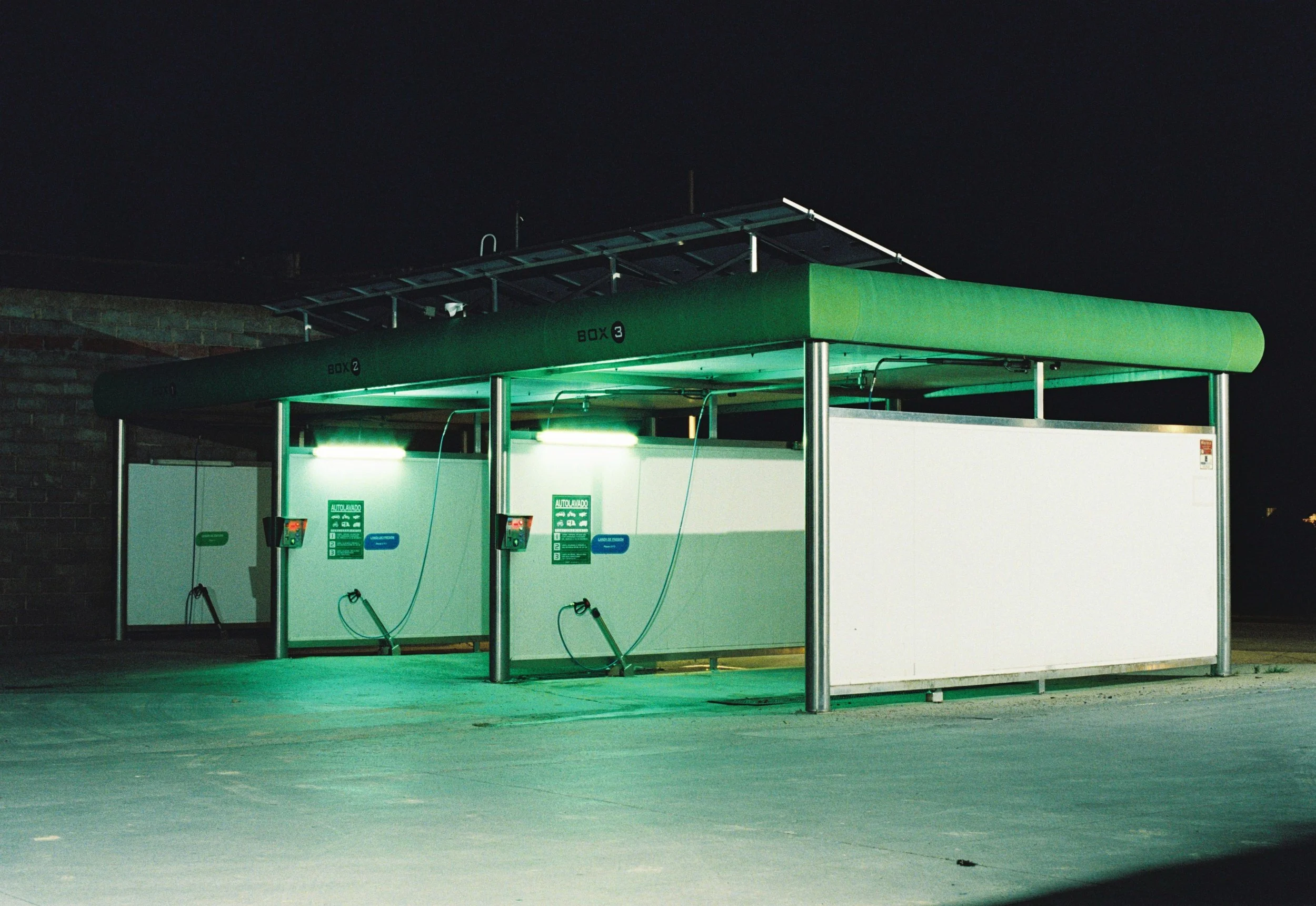 Estación de autoservicio de autoslavado con luces verdes, toldo verde y paredes blancas en la noche.
