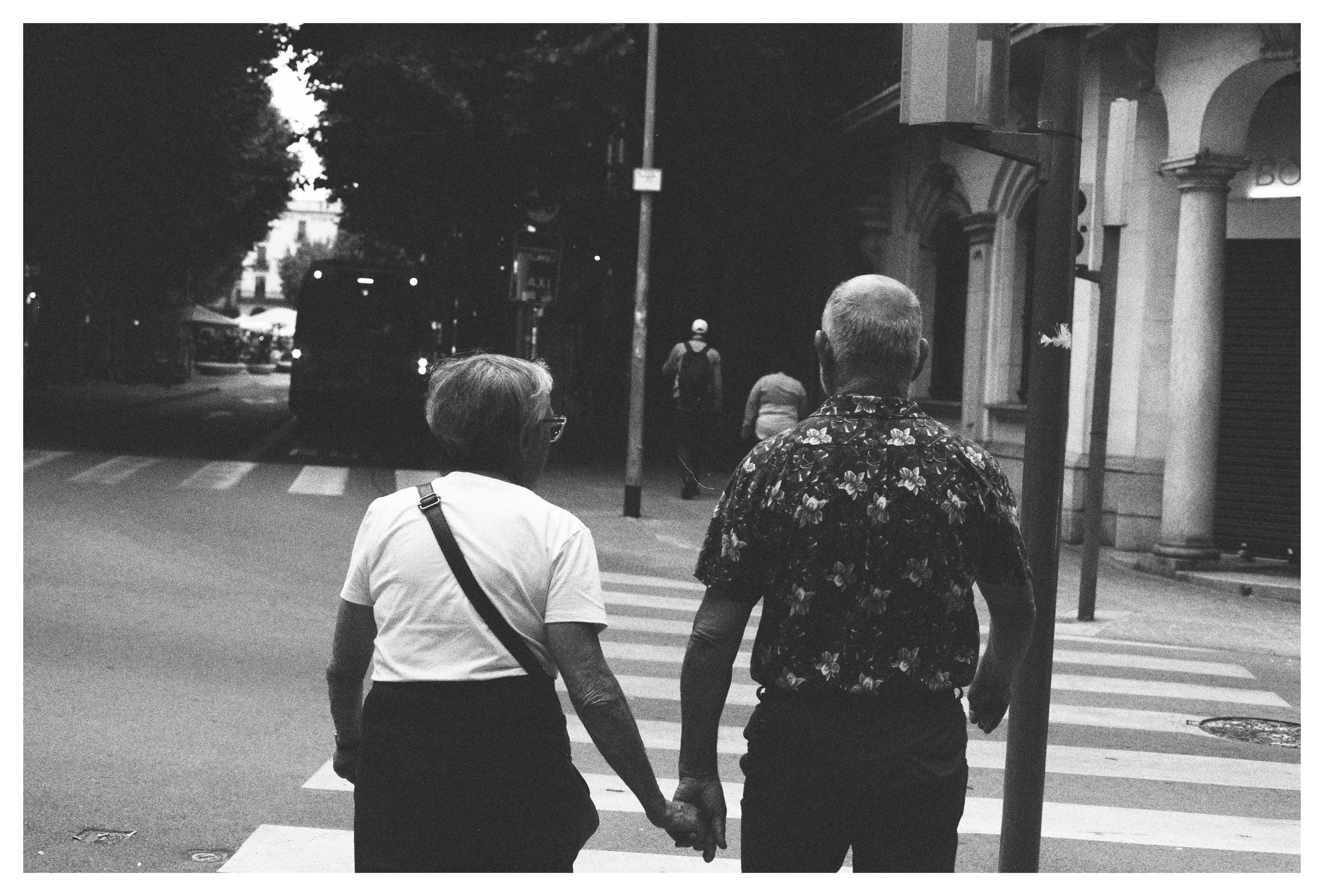 Una pareja de ancianos sosteniendo sus manos mientras cruzan la calle por un paso peatonal en una ciudad por la noche, en blanco y negro.