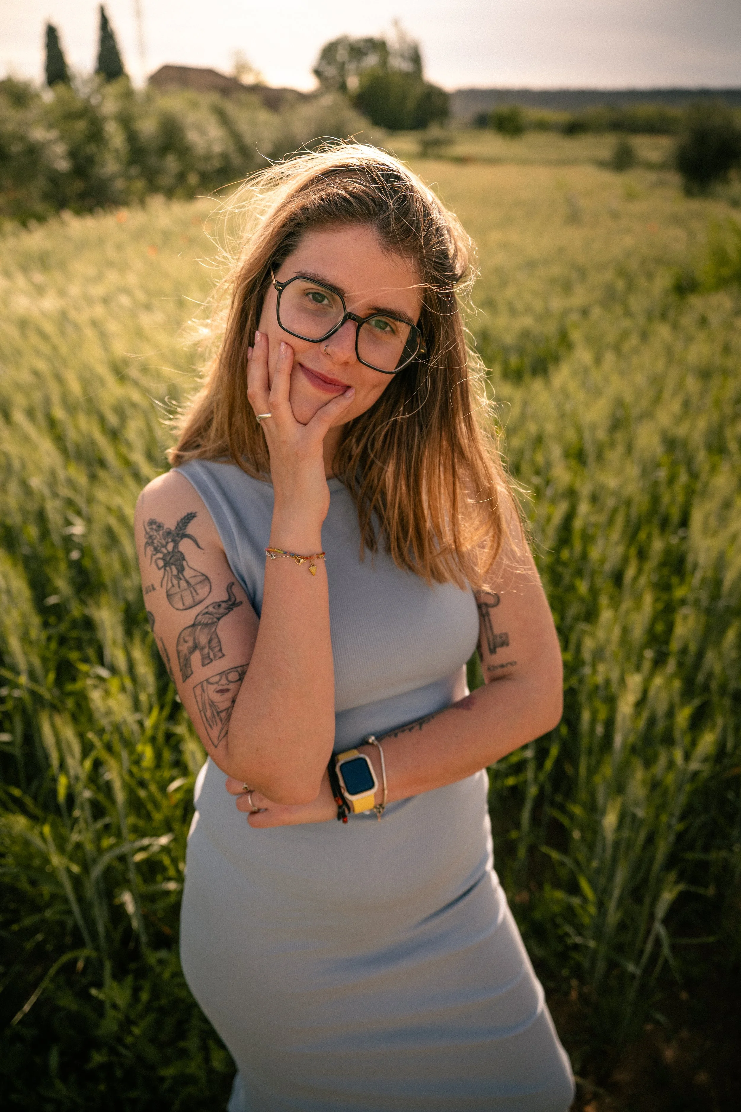 Joven mujer con gafas y tatuajes en un campo de césped, sonriendo y tocándose la mejilla con la mano izquierda, en un entorno natural durante el atardecer.